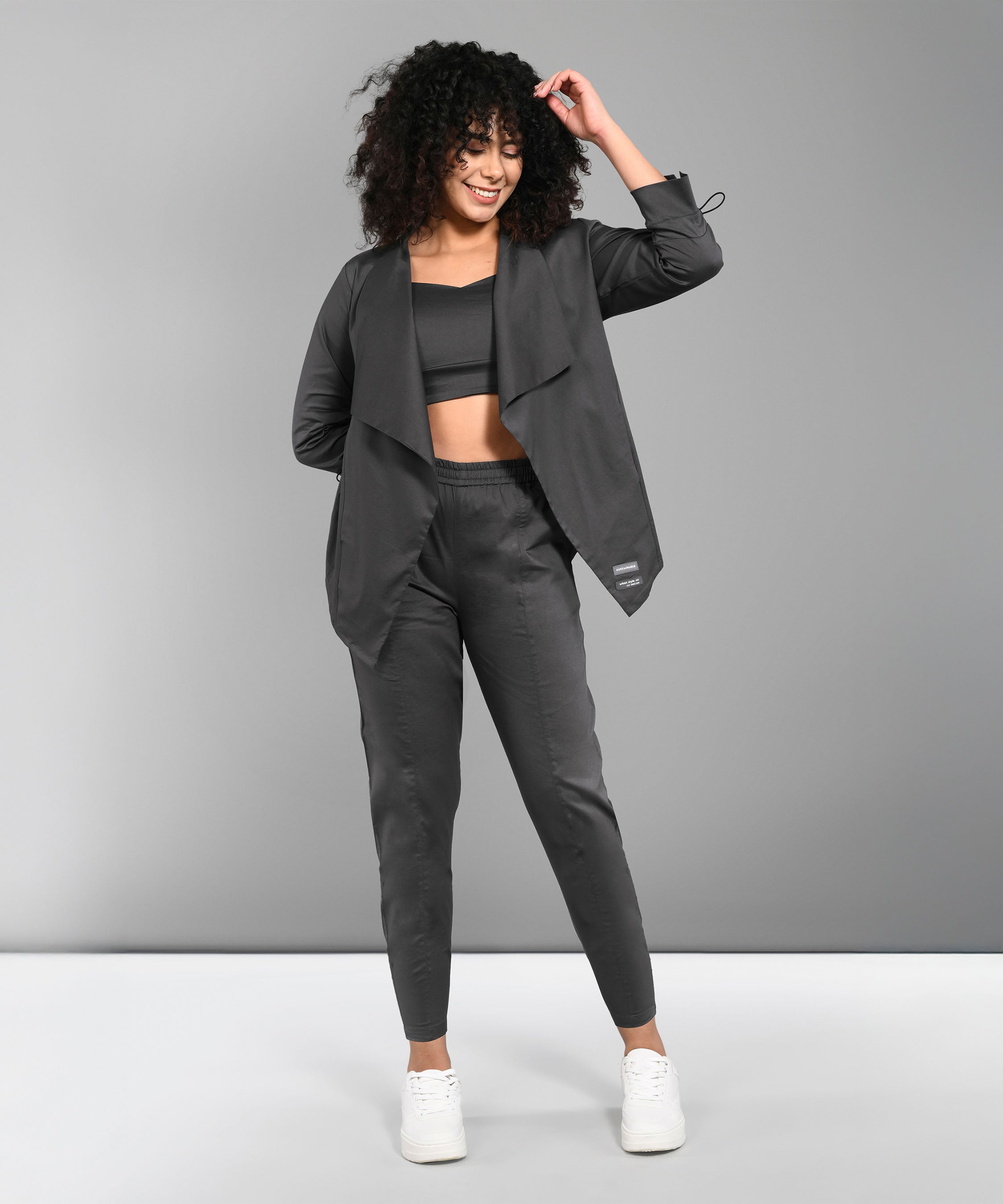 A woman with curly hair smiles and poses against a gray background, wearing the Timeless Brilliance Grey Co-ord Set with Crop Top and white sneakers.