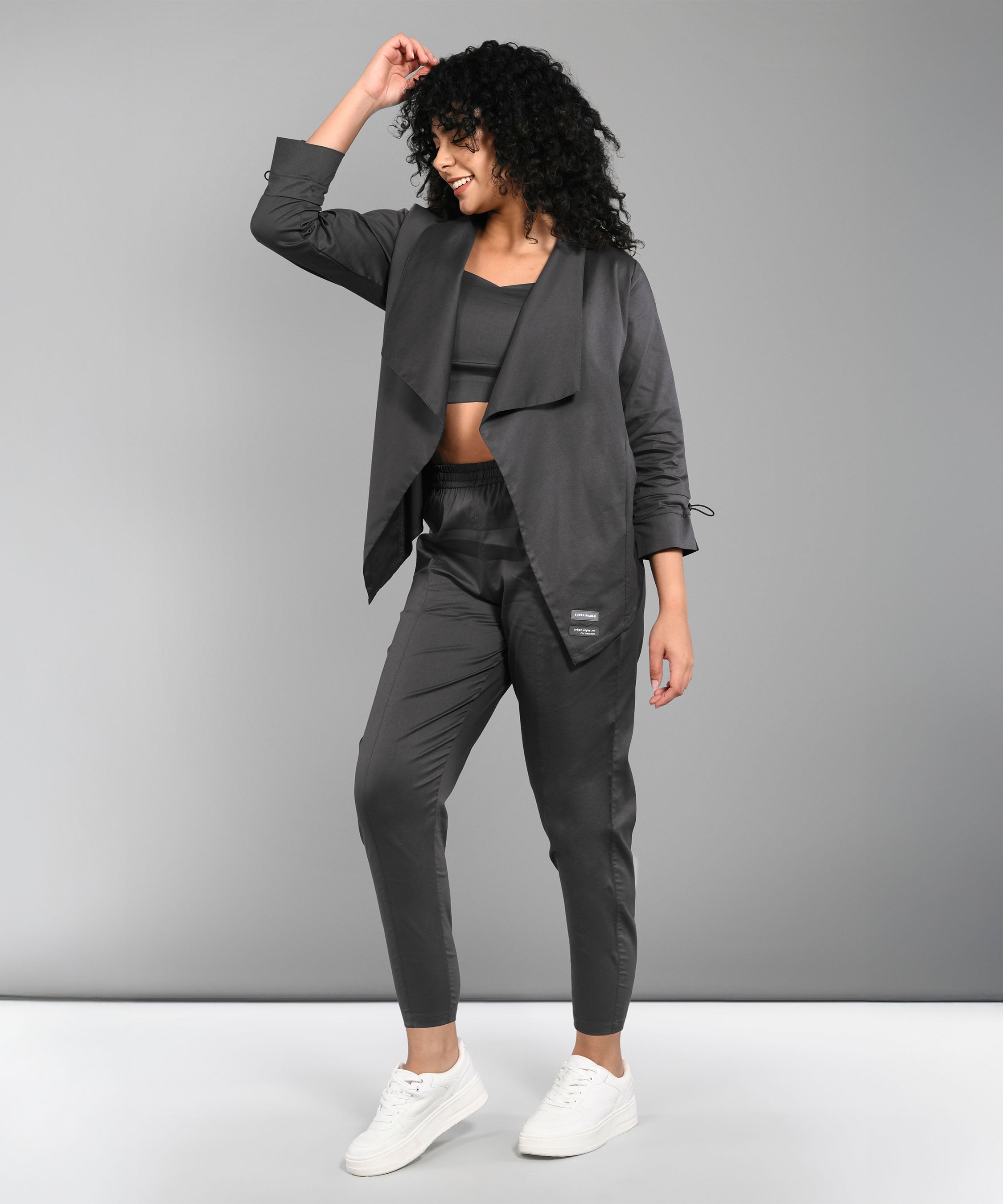 Model wearing the Timeless Brilliance Grey Co-ord Set with Crop Top, posing and smiling in white sneakers against a gray background.