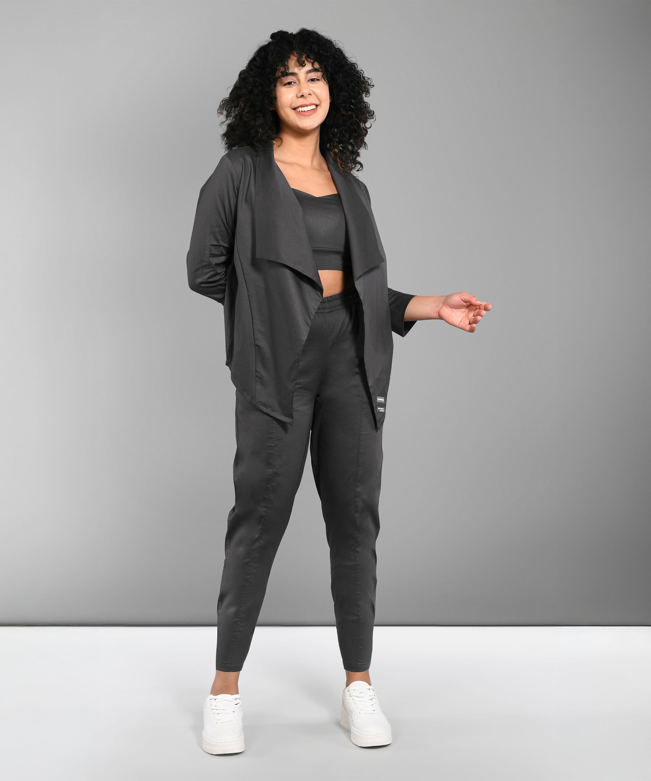 A woman with curly hair stands and smiles against a gray background, wearing the Timeless Brilliance Grey Co-ord Set with Crop Top and white sneakers.