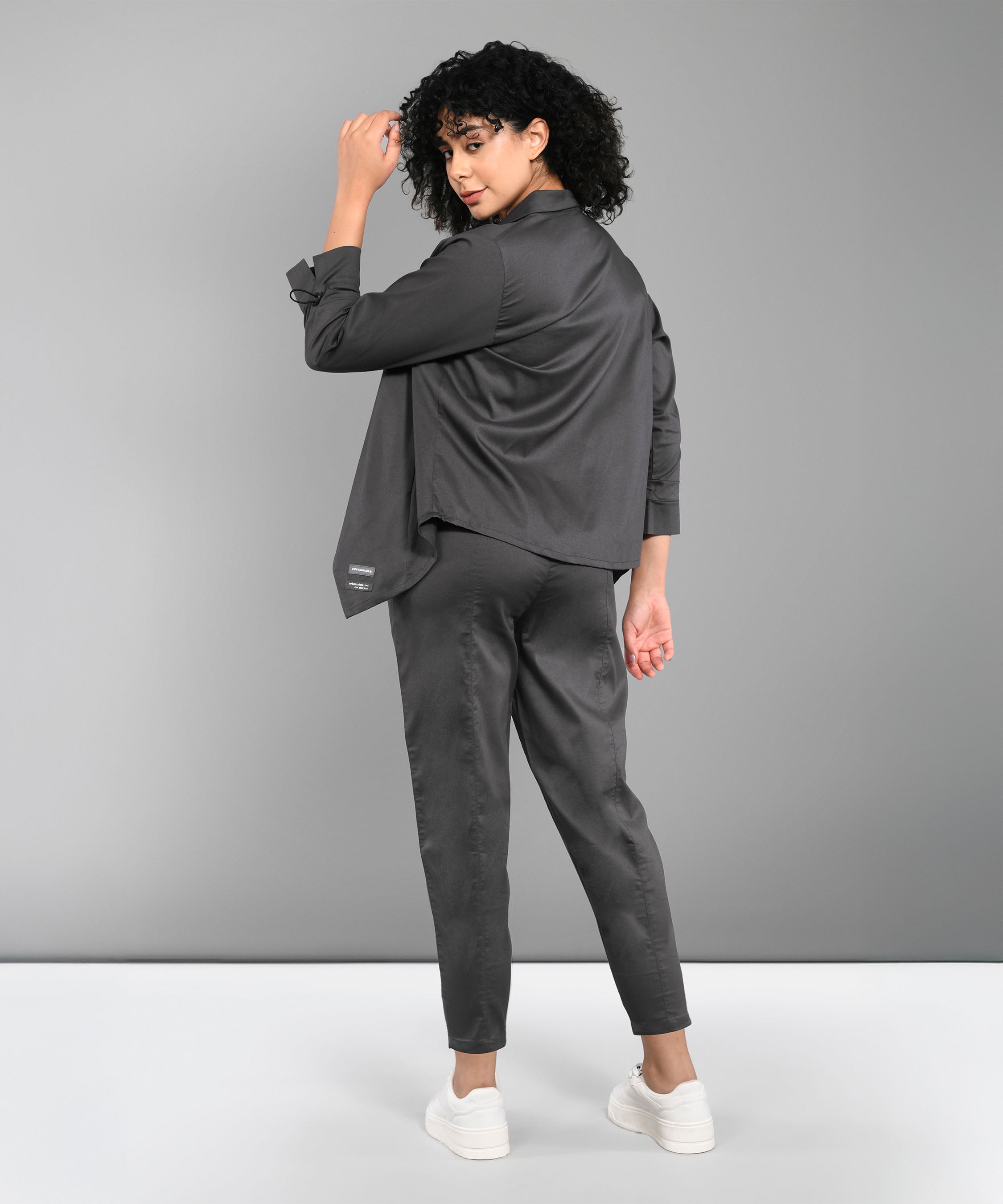A woman with curly hair stands and looks over her shoulder, wearing the Timeless Brilliance Grey Co-ord Set with Crop Top and white sneakers.