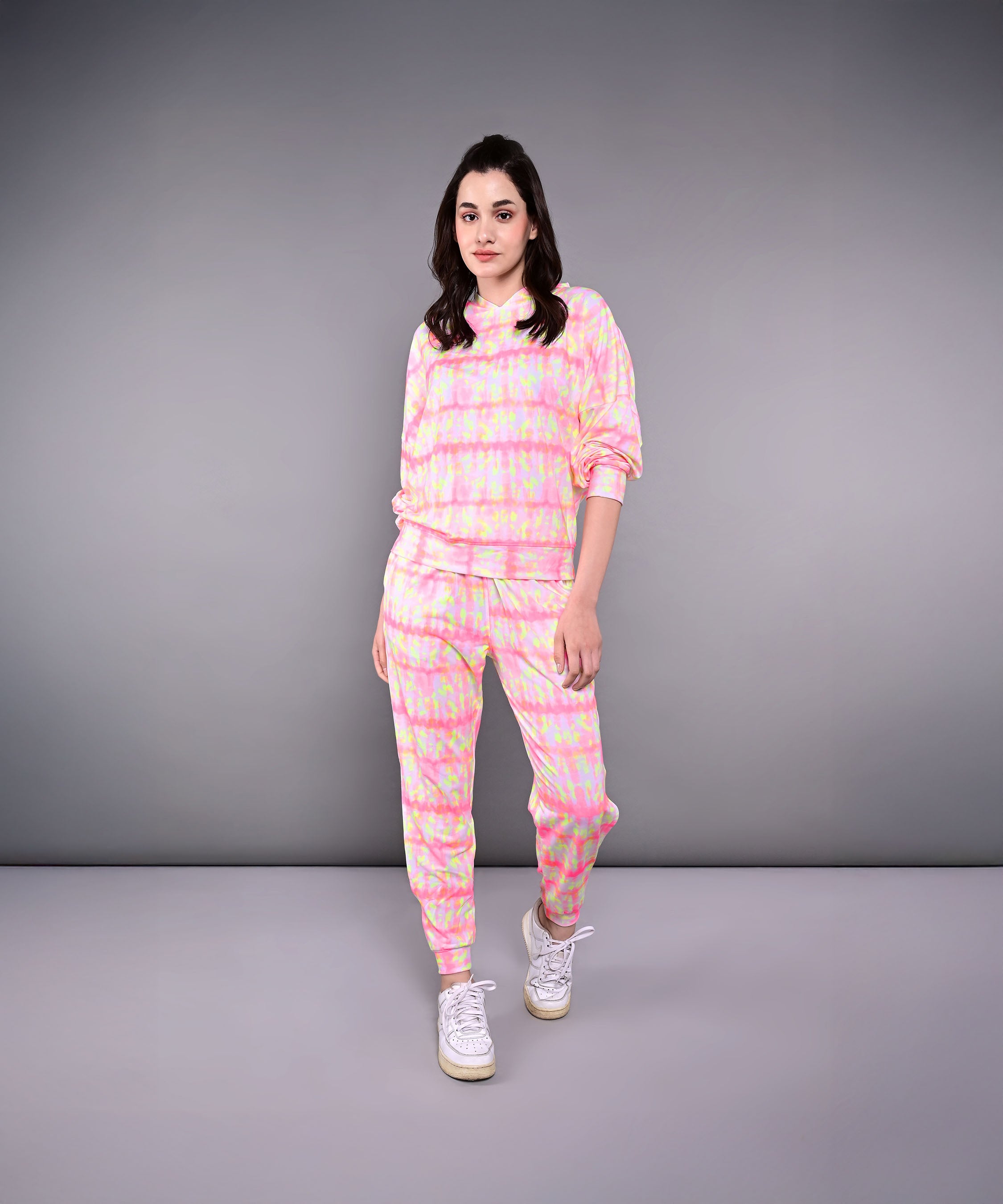 A woman models the Comfort Fit Tie-Dye Sweatshirt With Joggers Set, featuring a pink and yellow design in breathable poly-blend fabric, standing against a plain gray background.