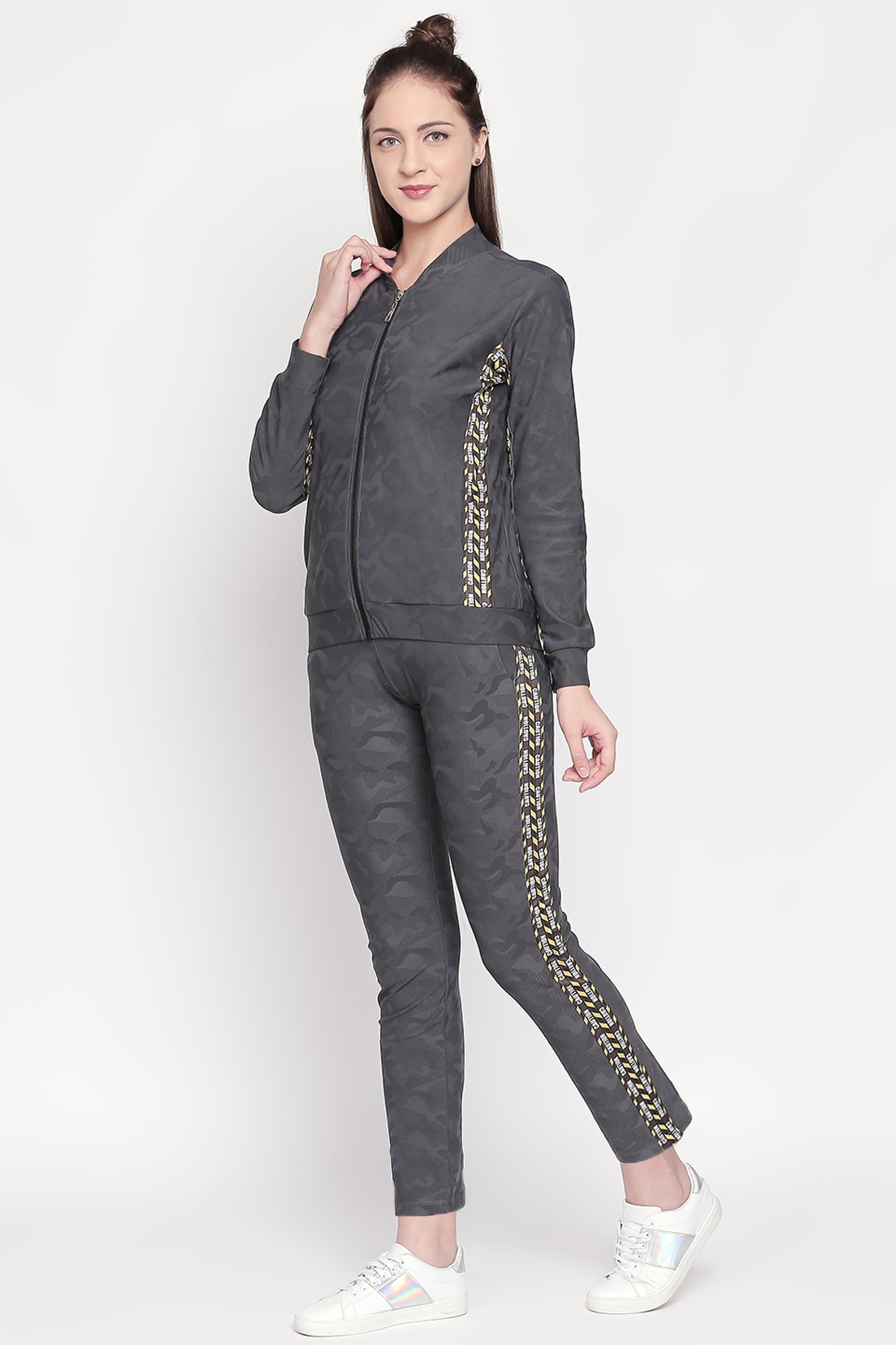 Woman smiling on a light background, wearing an Active Wear Printed Jacket in Grey from Tuna London. The dark gray jacket, featuring gold side stripes, is crafted from a comfortable polyester blend.