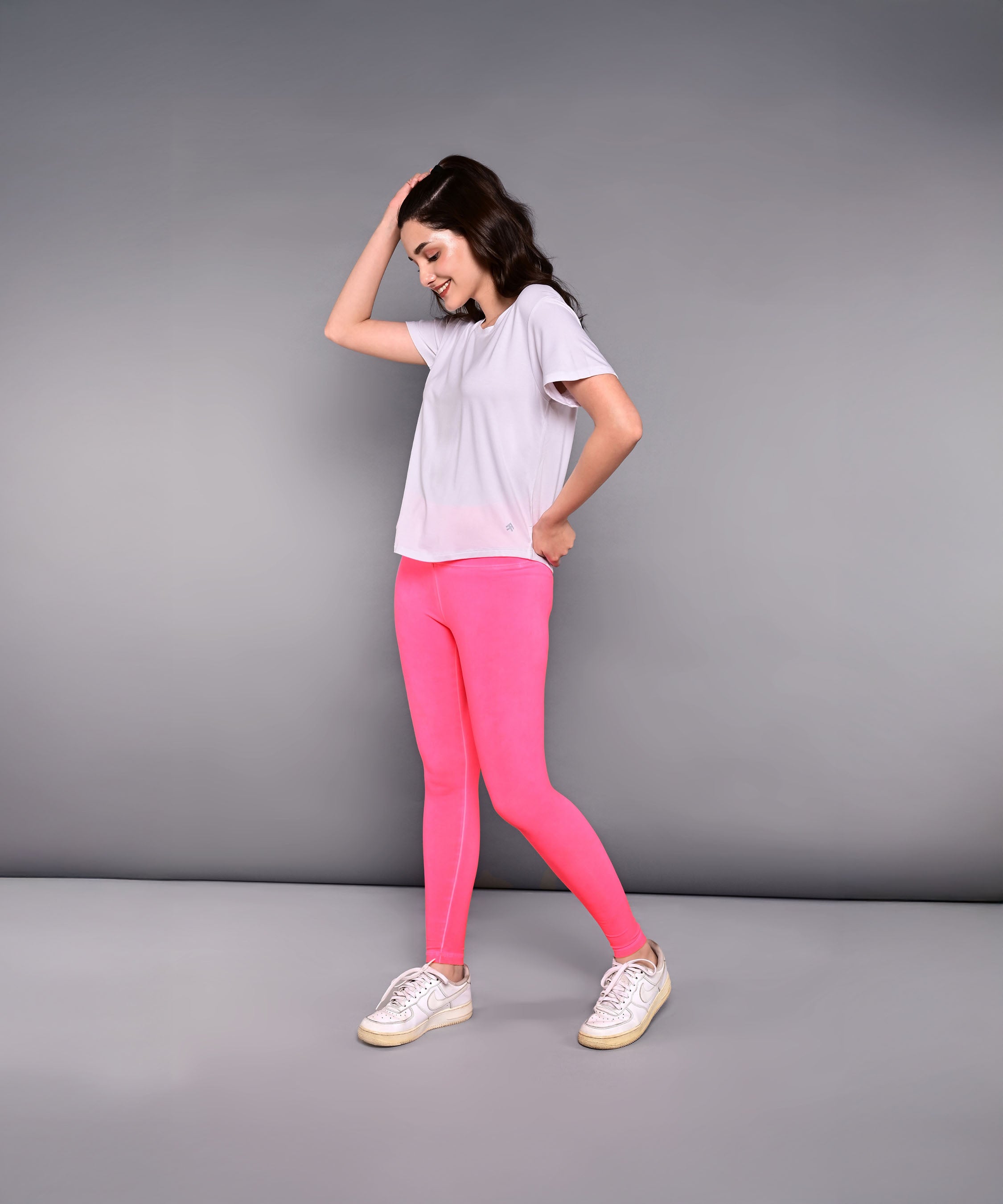 A woman smiles against a plain gray background, wearing the 'Pilates Has Your Back' printed tee in white with bright pink leggings.