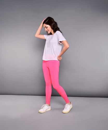 A woman smiles against a plain gray background, wearing the 'Pilates Has Your Back' printed tee in white with bright pink leggings.
