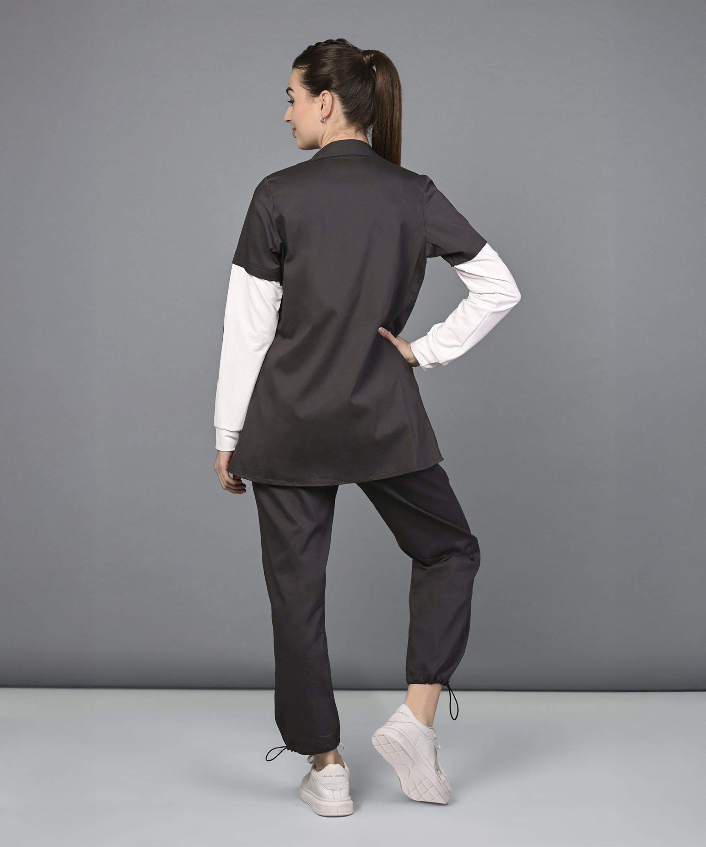 Woman in black Athleisure Blazer Tracks Two Piece Co-ord set and white sneakers stands with her back to the camera, hand on hip, against a gray backdrop—her style is effortlessly chic.