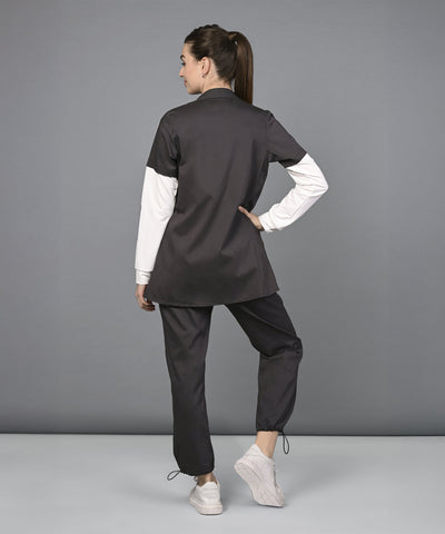 Woman in black Athleisure Blazer Tracks Two Piece Co-ord set and white sneakers stands with her back to the camera, hand on hip, against a gray backdrop—her style is effortlessly chic.