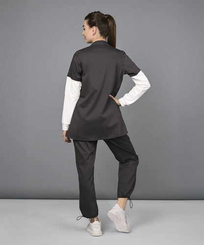 Woman in black Athleisure Blazer Tracks Two Piece Co-ord set and white sneakers stands with her back to the camera, hand on hip, against a gray backdrop—her style is effortlessly chic.