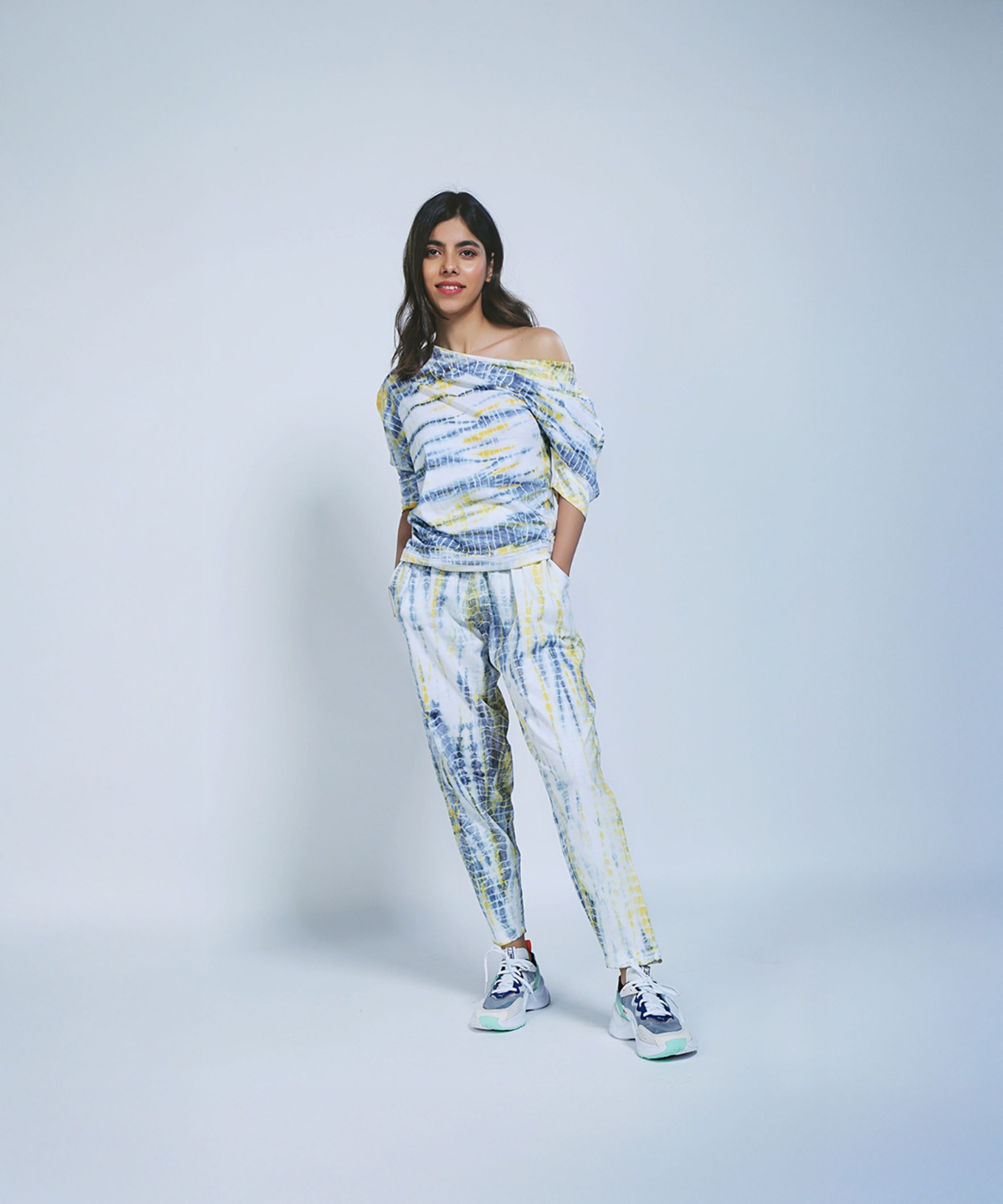 Woman wearing the One Side Off-Shoulder + Yoga Pants set in yellow & grey with sneakers, standing against a plain light background, hands in pockets.