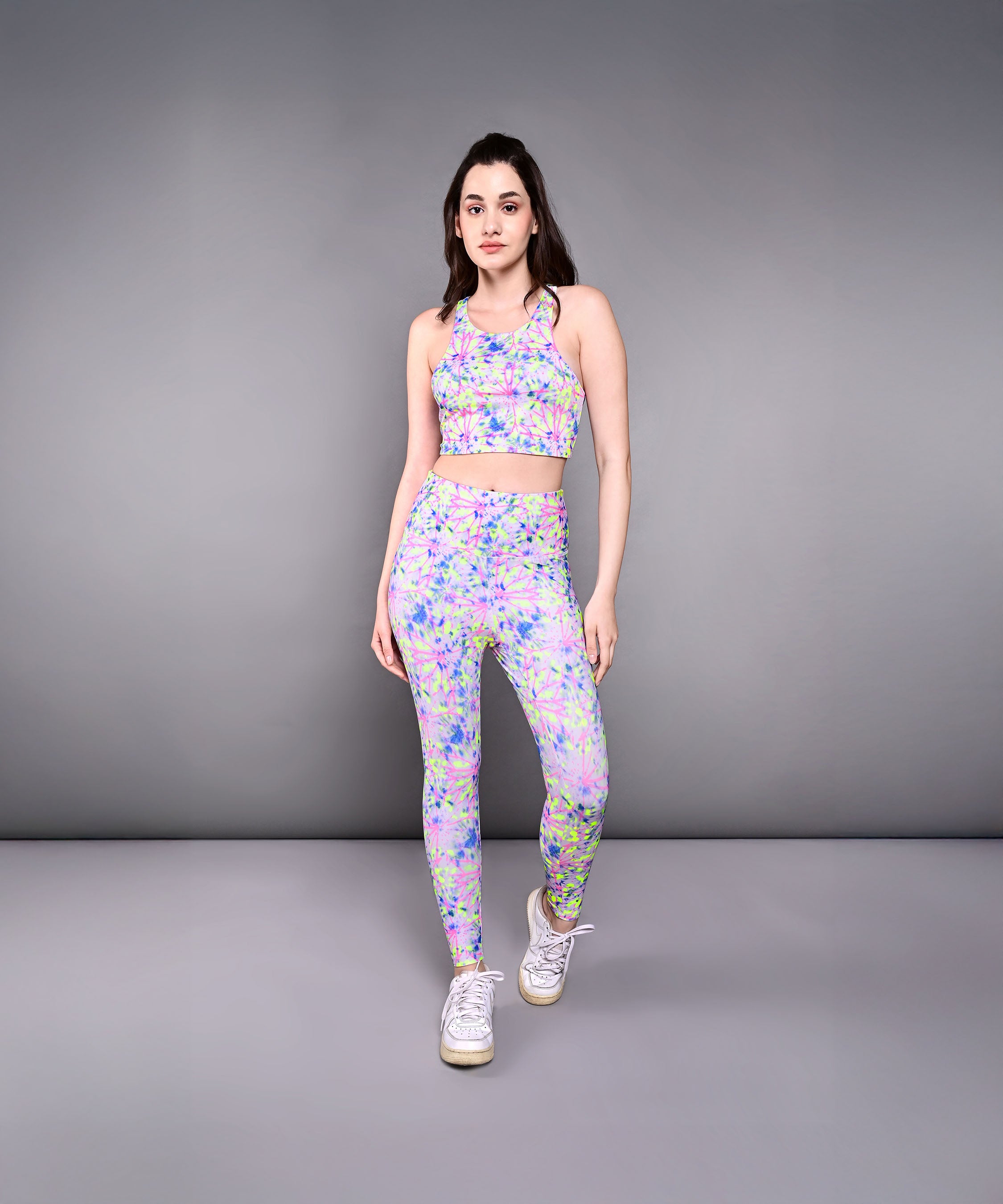A woman wearing Multicolour Neon Leggings and white sneakers stands against a plain gray background, highlighting the vibrant patterns and soft poly blend fabric of this activewear.