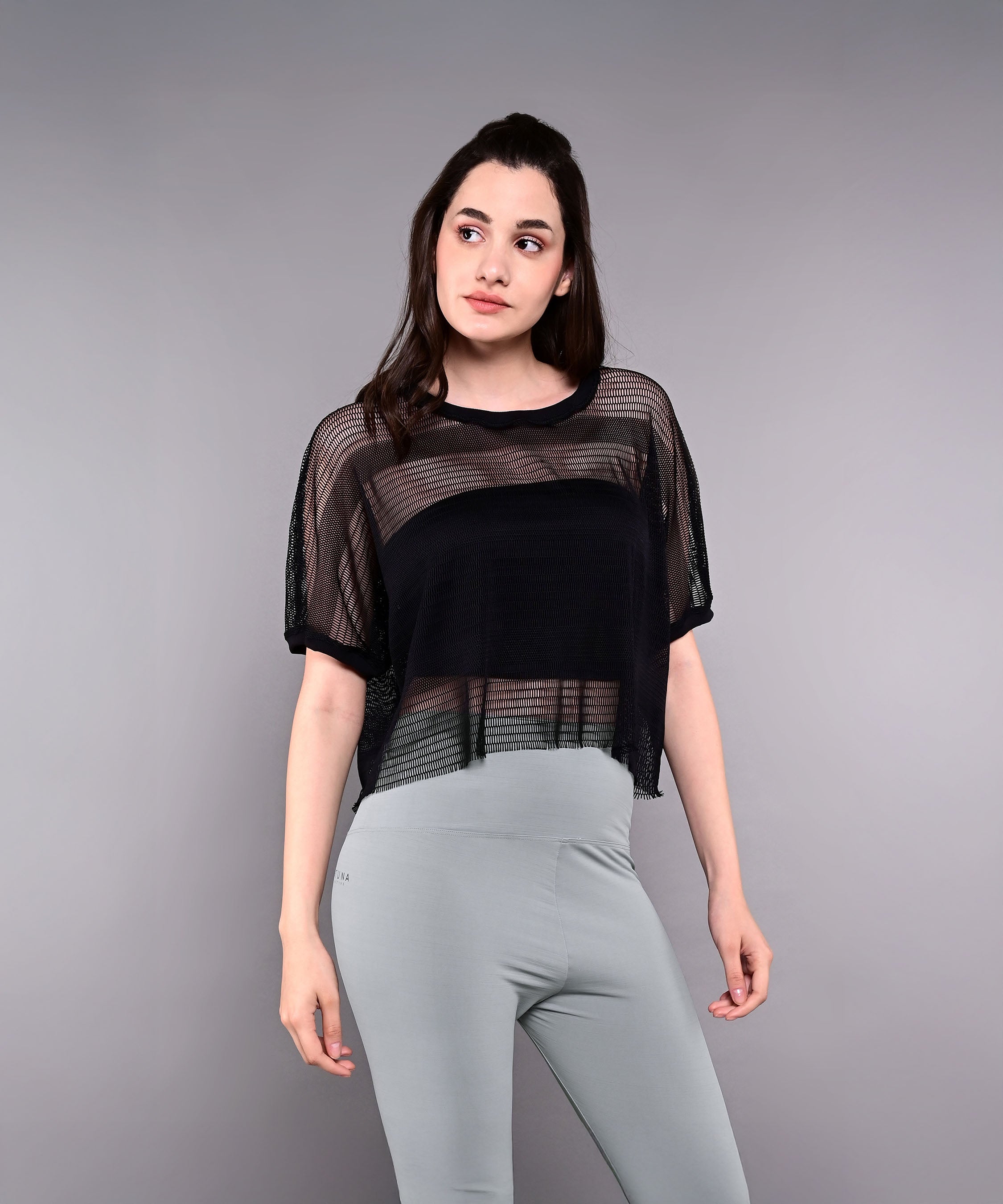 A woman wears the Tuna Active Black Netted Top layered over a black tank with light gray leggings, standing against a plain gray background.