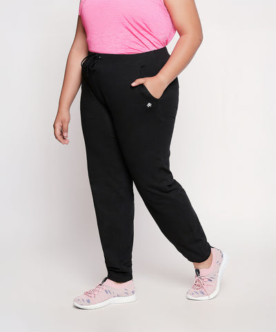 Woman in a pink top, Classic Joggers - Plus Size 02 (black high-stretchy workout pants), and pink shoes stands with one hand in her pocket.