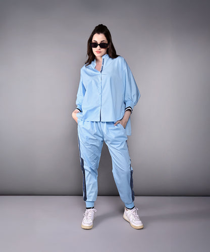 Woman wearing the Denim Accented Set—a light blue 100% cotton batwing sleeve shirt and joggers—stands with hands in pockets and sunglasses against a plain gray background.