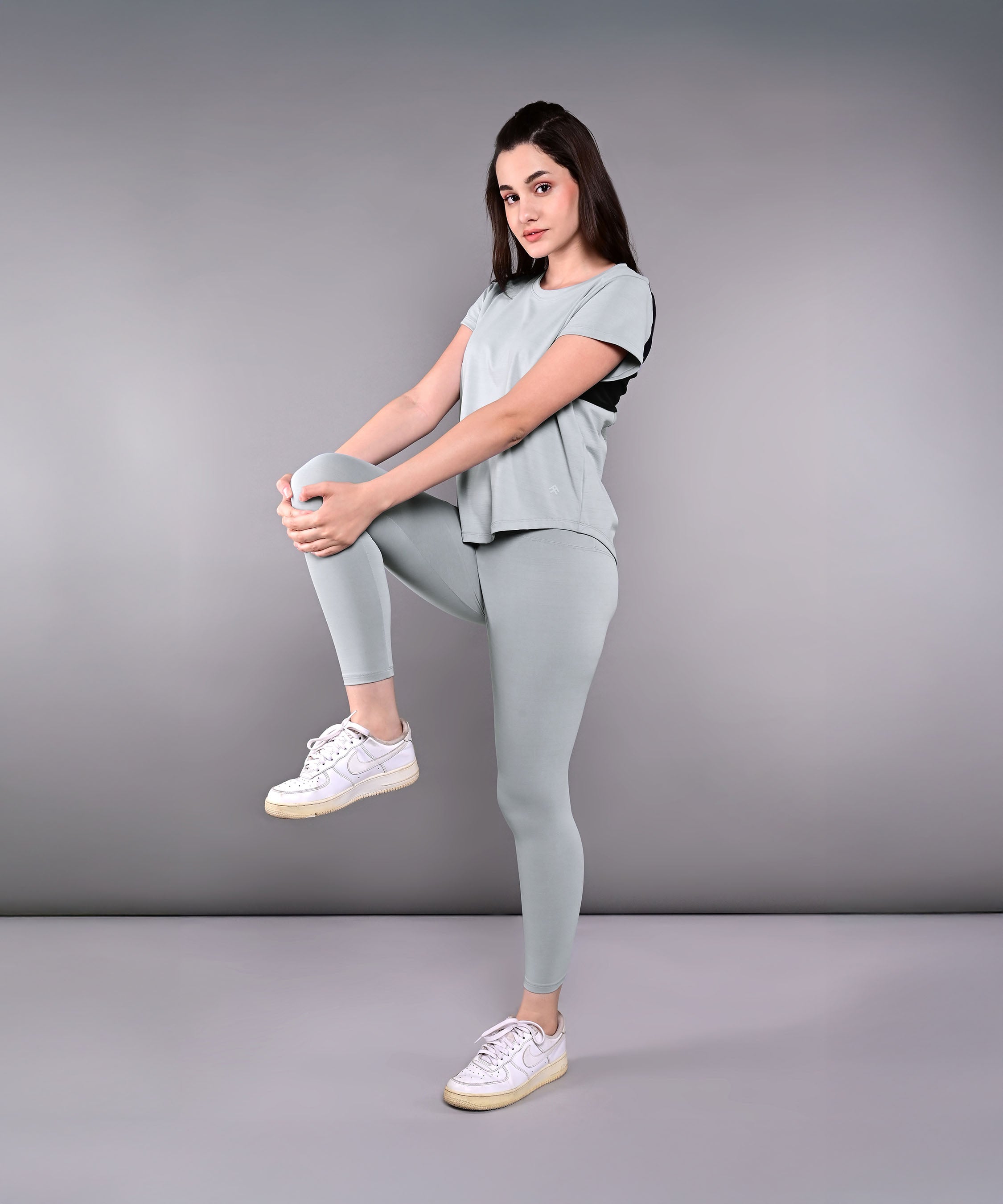 A woman showcases the Dynamic Grey Performance Set, standing on one leg and holding her raised knee against a plain gray background.