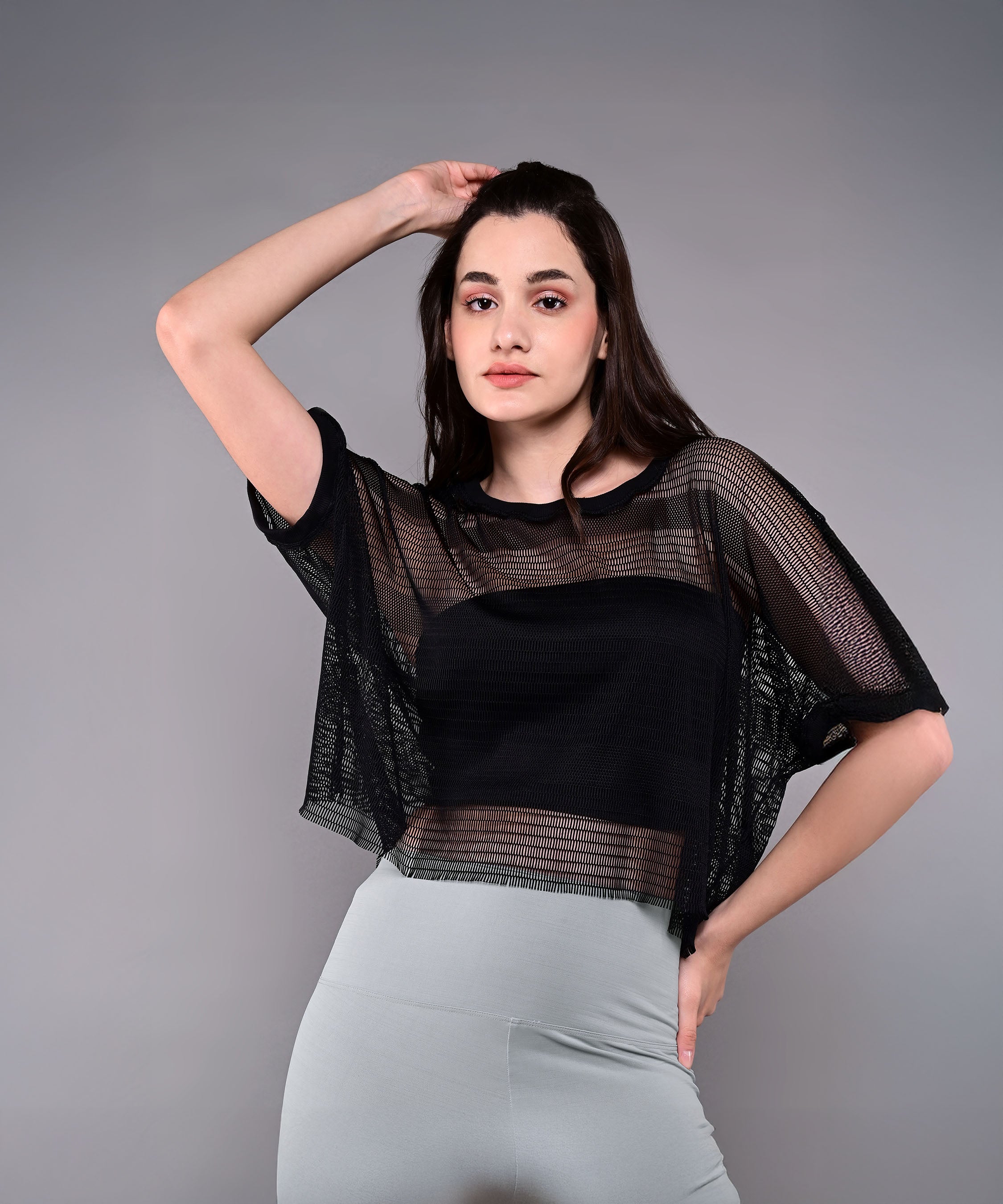A woman poses in the Tuna Active Black Netted Top with light gray pants, set against a plain gray background.