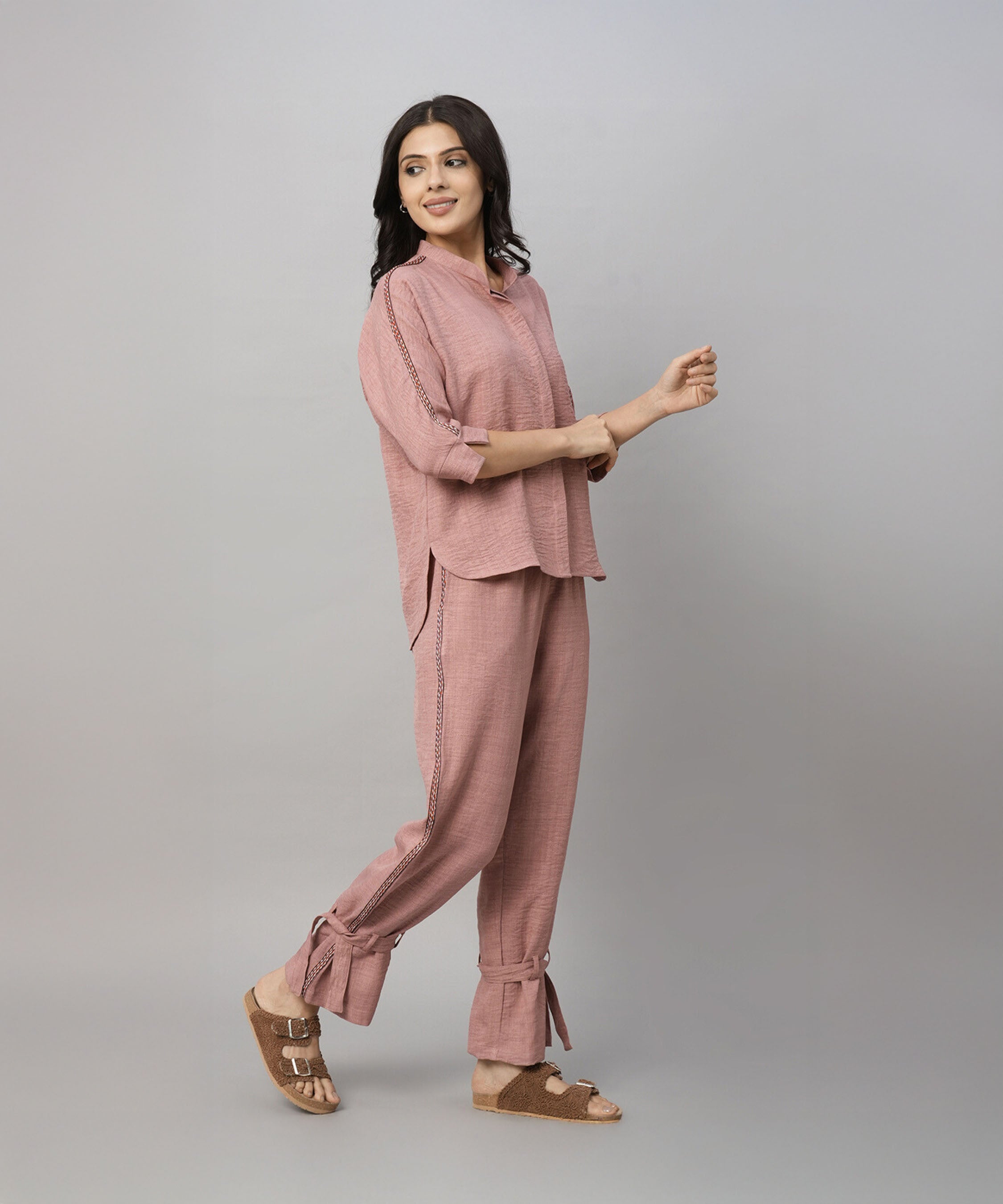 A woman wearing the Vintage Pink Loose Shirt and Tie Up Pant Cord Set made from 100% cotton, paired with brown sandals, stands smiling against a plain gray background.
