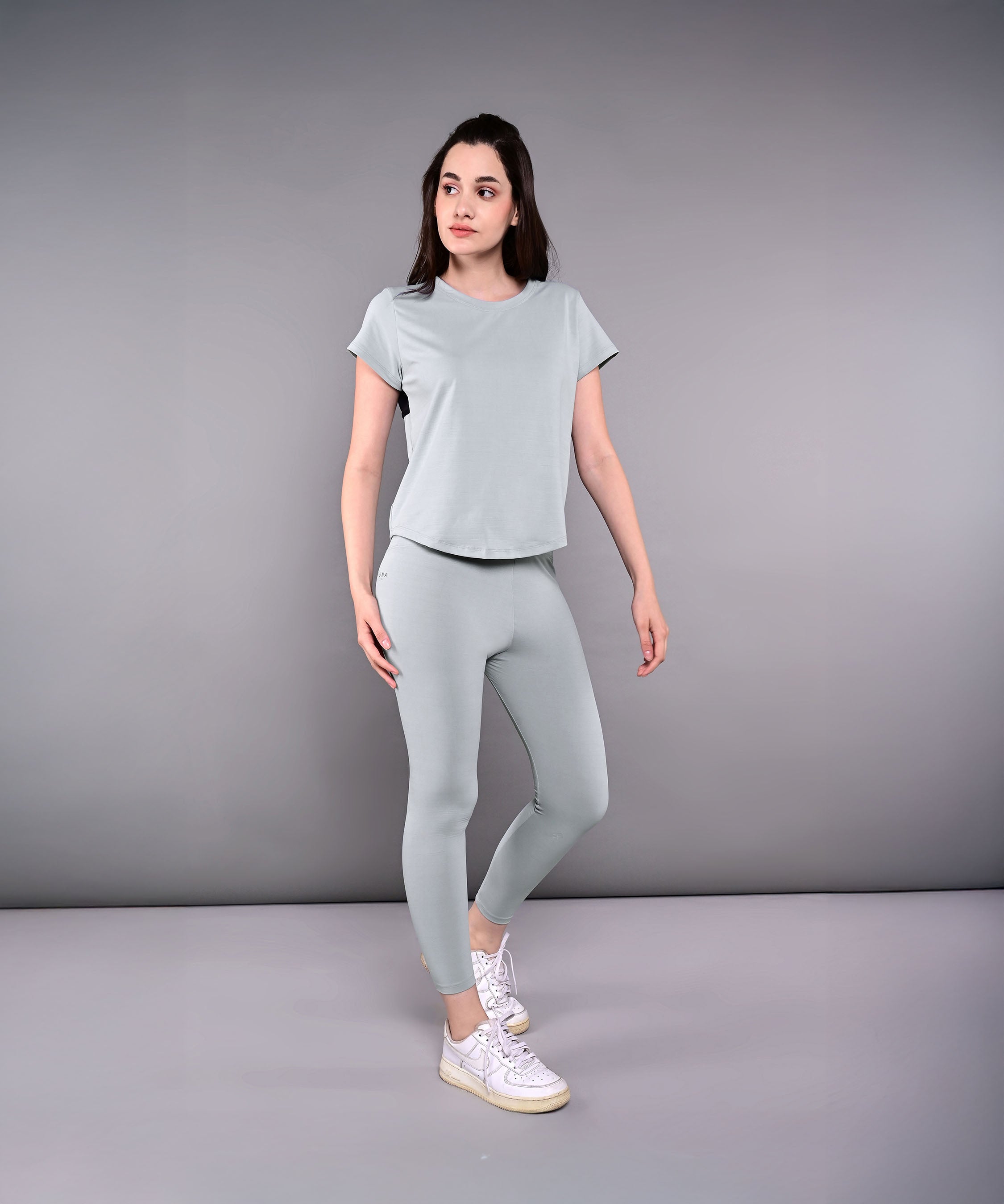A woman stands in a studio wearing the Dynamic Grey Performance Set—athletic leggings and a grey racerback top with moisture-wicking properties—against a neutral background.