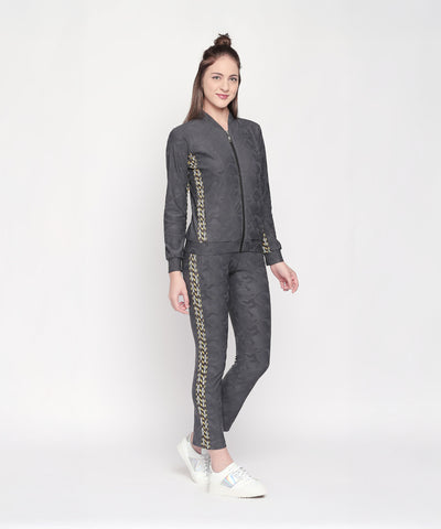 A woman models Self Printed Grey TrackPants—dark gray with gold patterns—smiling confidently against a plain white background, highlighting their stylish and comfortable athleisure appeal.