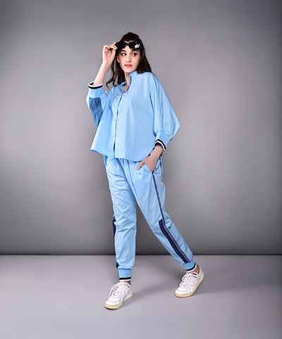 Woman models the Denim Accented Set—light blue shirt and joggers paired with white sneakers—against a plain gray background, highlighting the comfort of 100% cotton.