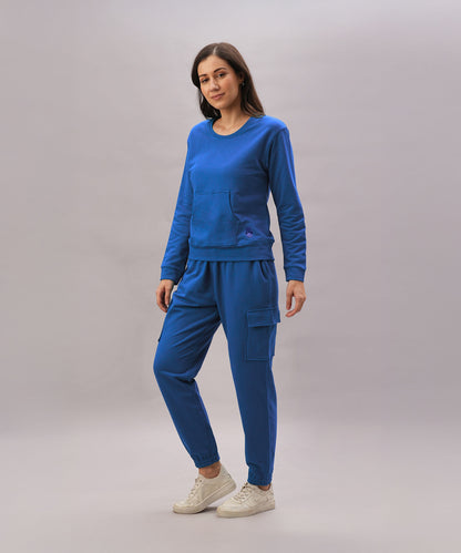 A woman stands against a plain background in the Soft Royal Blue Trackset, featuring cargo-style joggers and white sneakers, crafted from a breathable cotton-viscose blend.