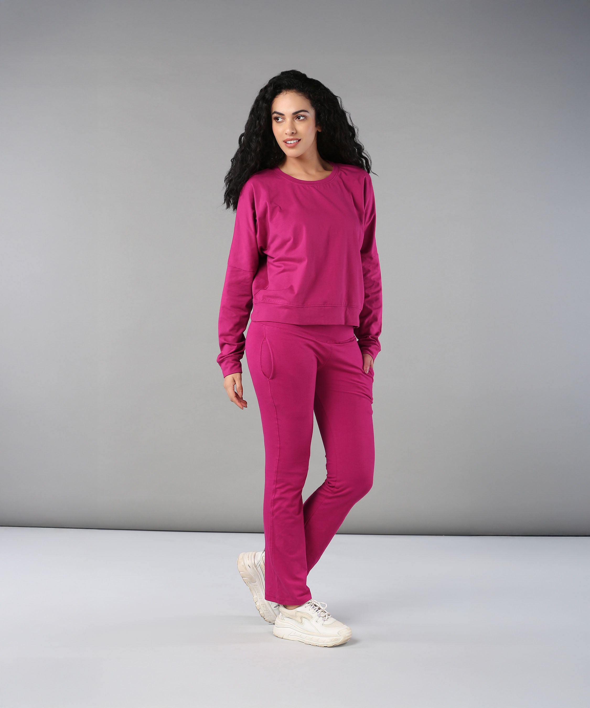 Woman with curly hair wears the Deep Pink Comfort Duo tracksuit and white sneakers, standing against a gray background.