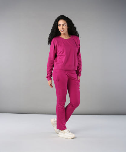 Woman with curly hair wears the Deep Pink Comfort Duo tracksuit and white sneakers, standing against a gray background.