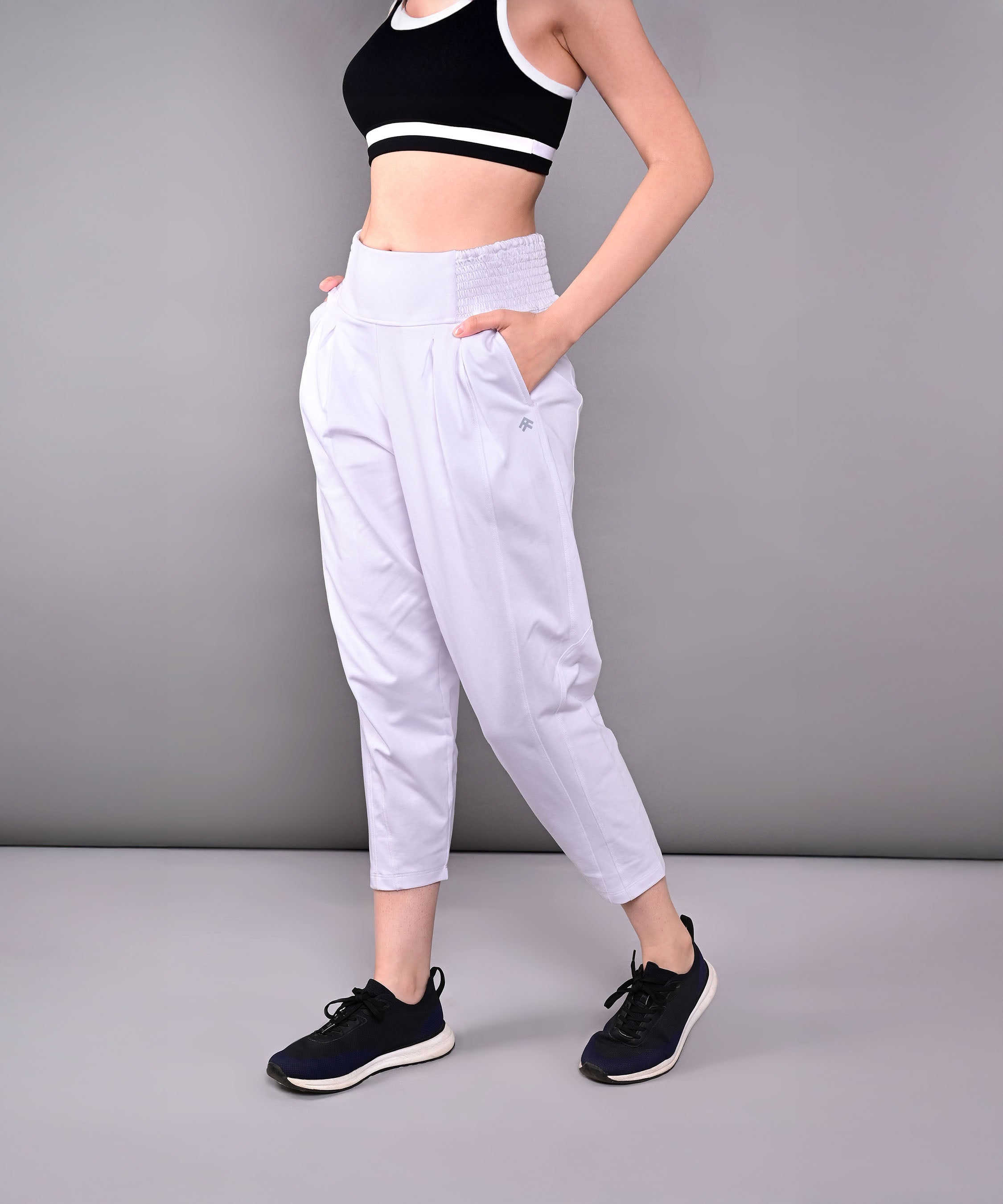 Woman wearing a black sports bra, black sneakers, and Pleated Yoga Pant With Smocked Waistband in white against a grey background.