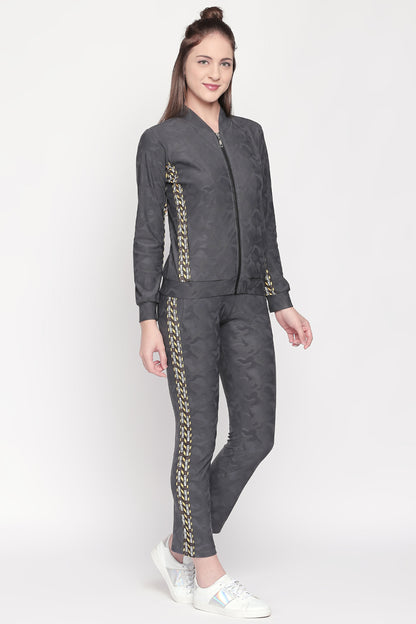 A woman wearing a dark polyester blend tracksuit with patterned stripes, white sneakers, and the Active Wear Printed Jacket in Grey from Tuna London stands and smiles.