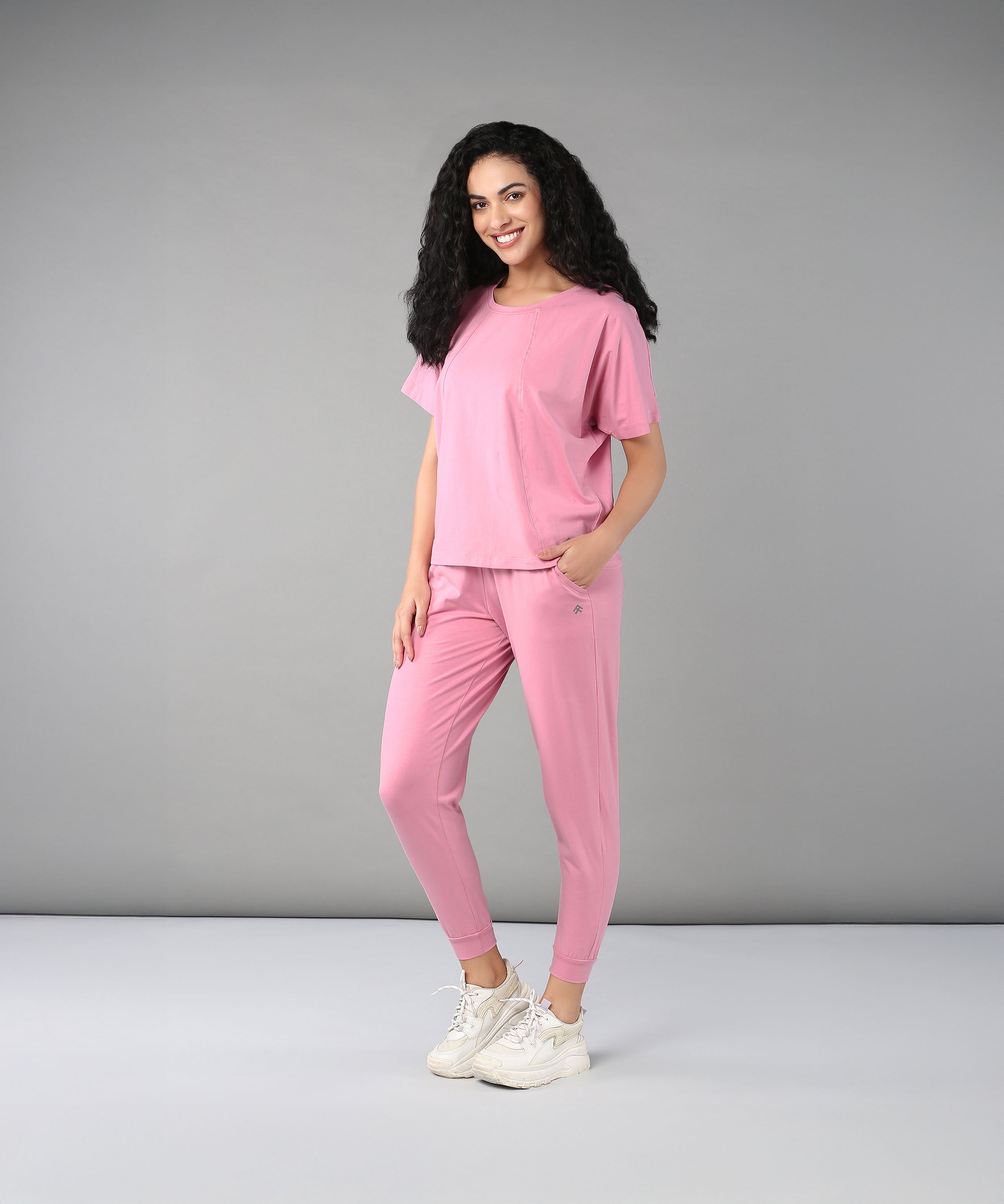 A woman with curly hair stands and smiles in a studio, wearing the Flamingo Pink Chic Ensemble—a racer back top made from buttery soft fabric paired with Cotton Lycra joggers.