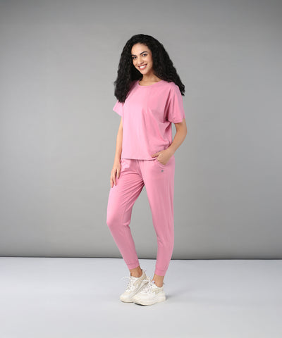 A woman with curly hair stands and smiles in a studio, wearing the Flamingo Pink Chic Ensemble—a racer back top made from buttery soft fabric paired with Cotton Lycra joggers.