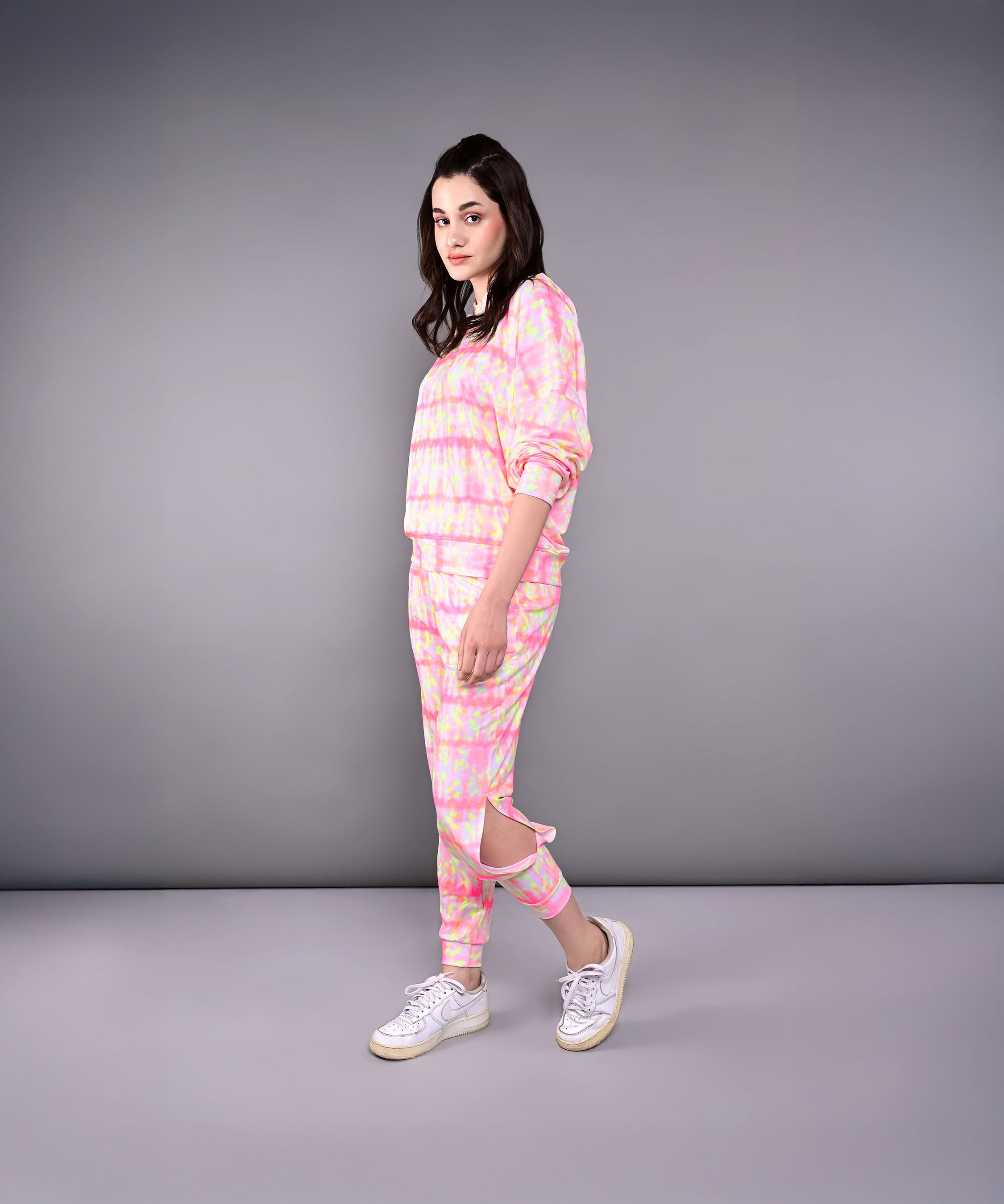 A woman models the Comfort Fit Tie-Dye Sweatshirt With Joggers Set, featuring a pastel design and one jogger pant leg cut and hanging, against a gray background.