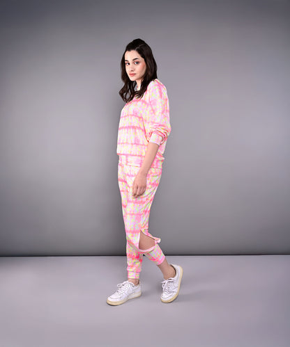 A woman models the Comfort Fit Tie-Dye Sweatshirt With Joggers Set, featuring a pastel design and one jogger pant leg cut and hanging, against a gray background.