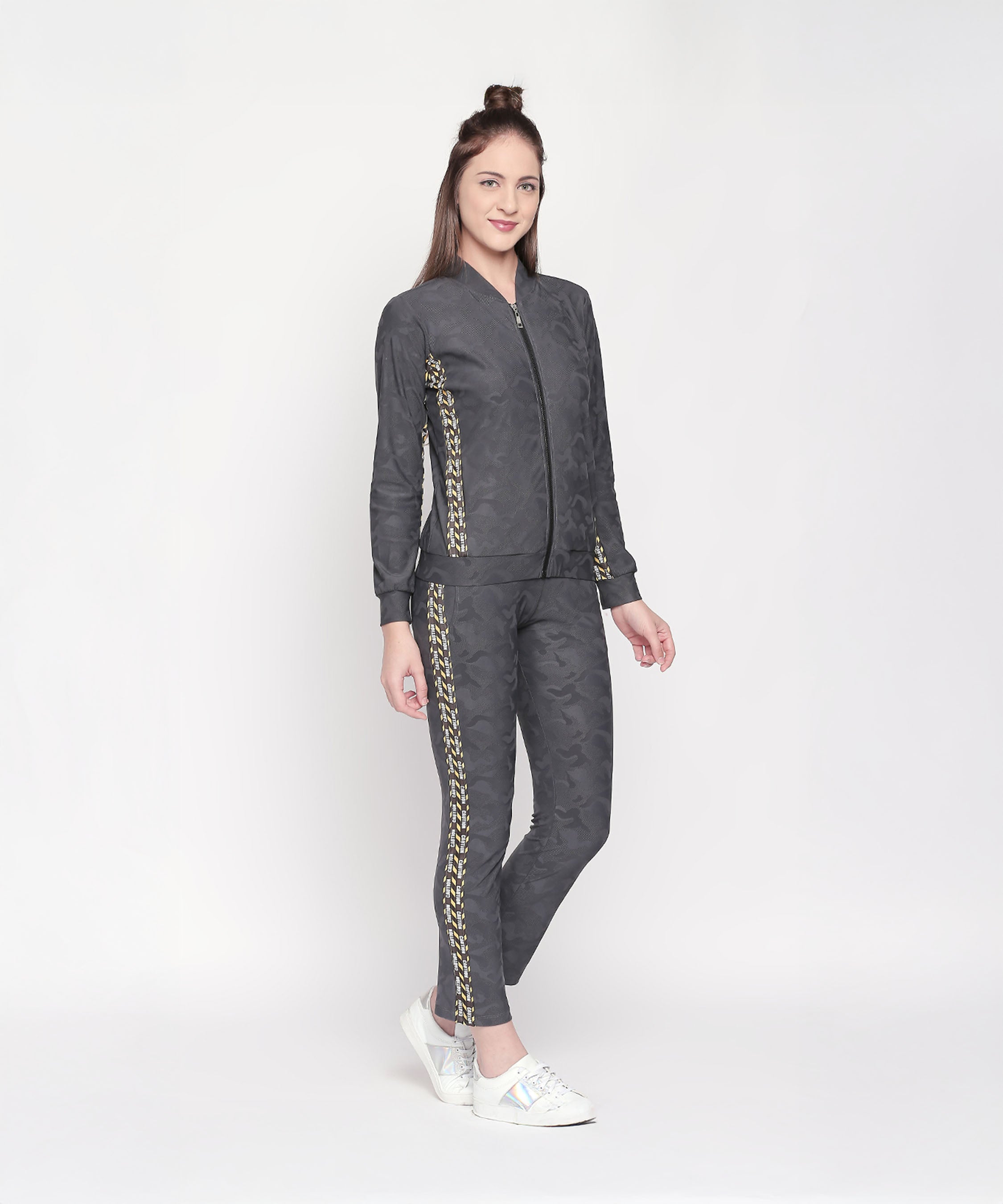 A woman smiles against a white background, wearing the Camouflage Print Tracksuit featuring a V-neck collar jacket and elasticated track pants with yellow patterns, paired with white sneakers.