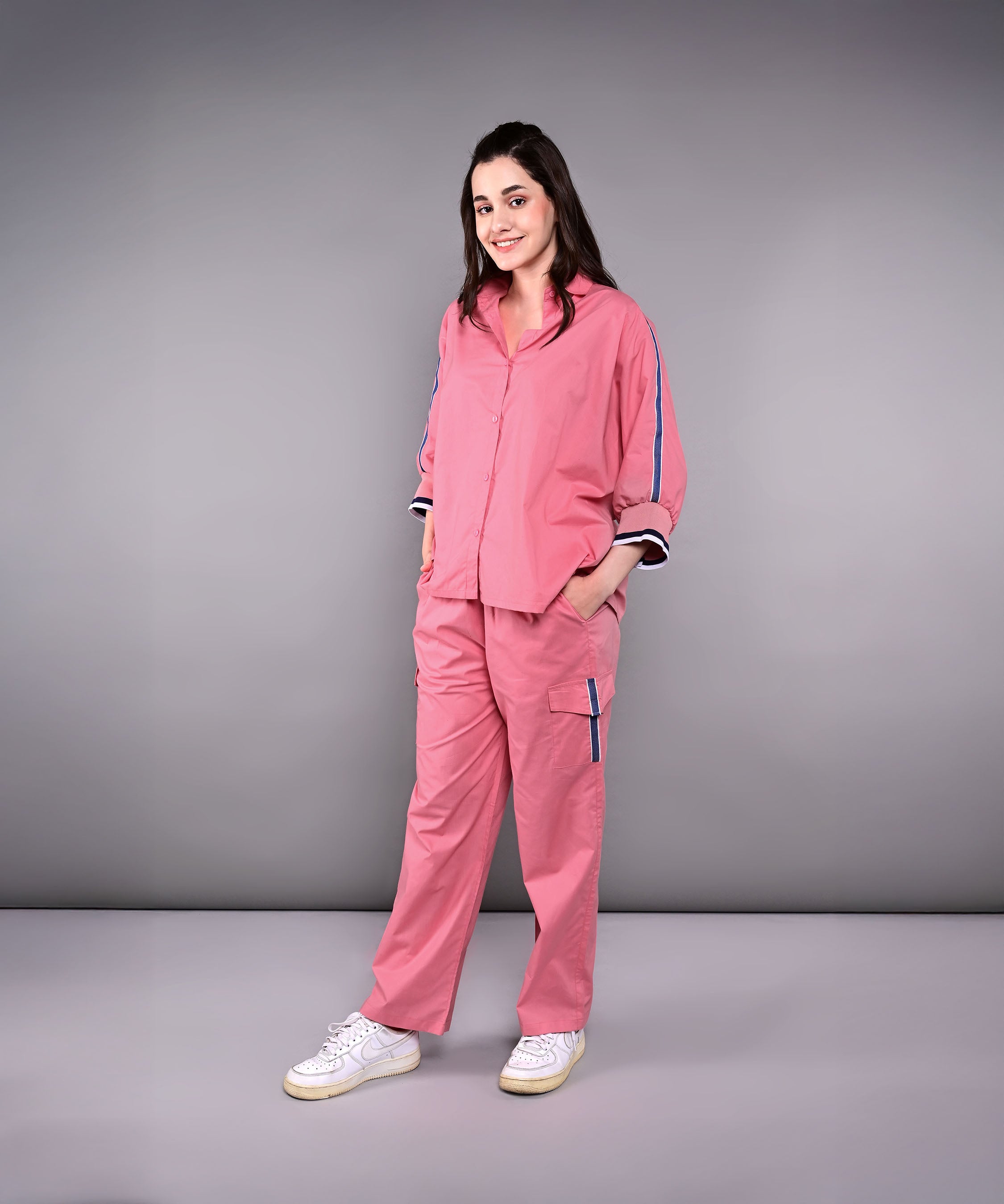 Woman wearing the Effortlessly Chic Set—a pink cotton co-ord with cargo track pants—smiling with hands in pockets, posed against a gray background.