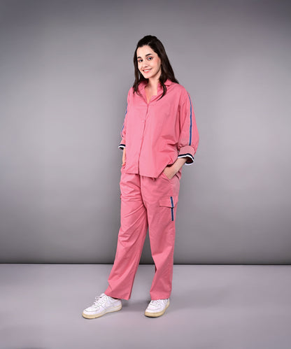 Woman wearing the Effortlessly Chic Set—a pink cotton co-ord with cargo track pants—smiling with hands in pockets, posed against a gray background.