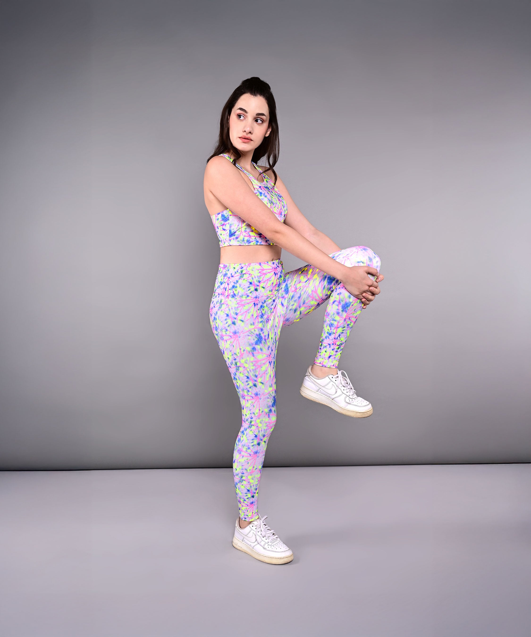 Woman in colorful activewear stands on one leg, holding her knee to her chest, showcasing Multicolour Neon Leggings against a gray background.