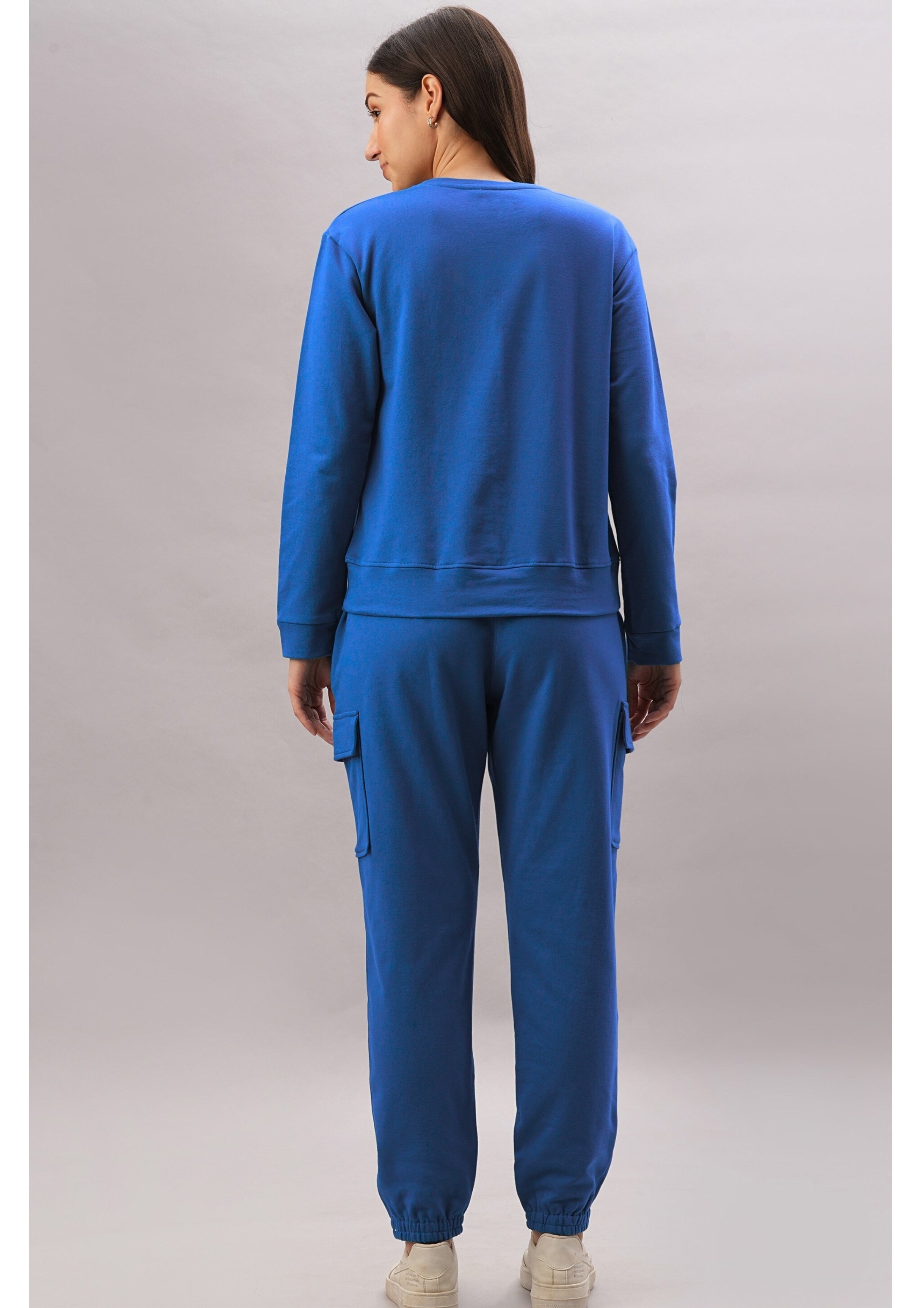 A woman wears the Soft Royal Blue Trackset with cargo-style joggers, standing with her back to the camera against a plain backdrop.