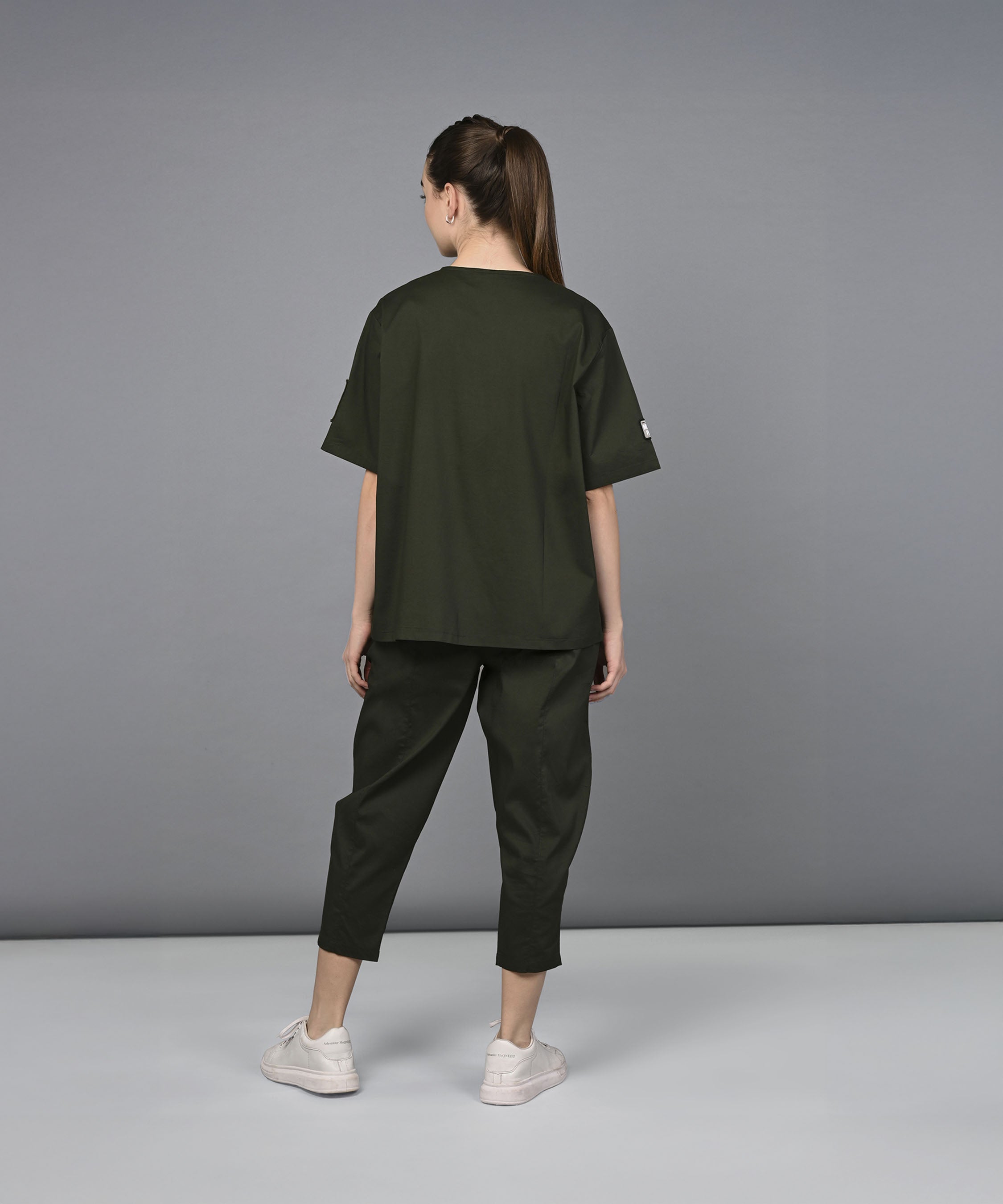 A woman wearing the Military Green Open Jacket & Yoga Pants with Bustiere set and white sneakers stands facing away against a plain gray background.