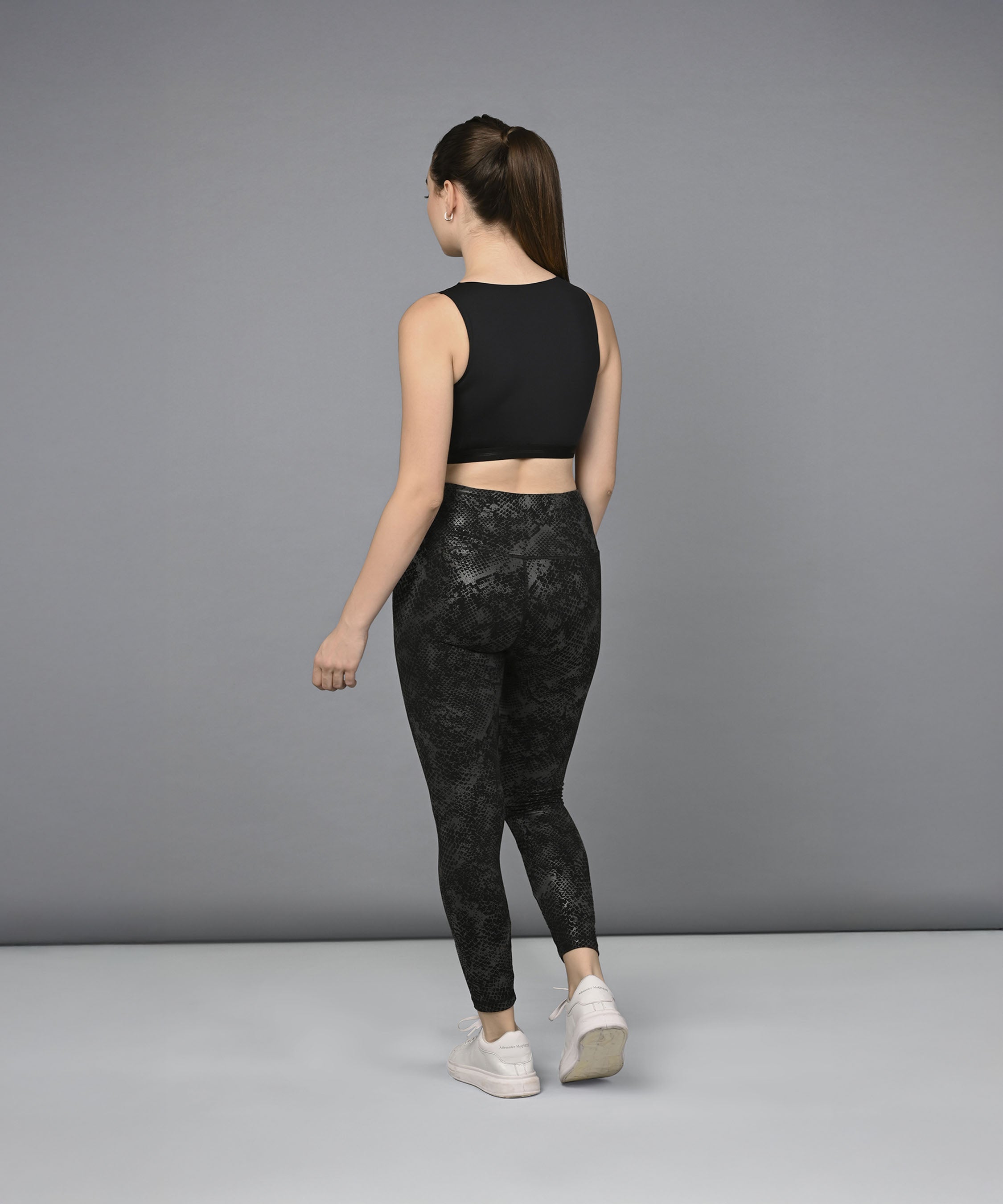 A woman models the Zipped Sports Bra & Metallic Legging set in black, standing with her back to the camera against a plain gray background.