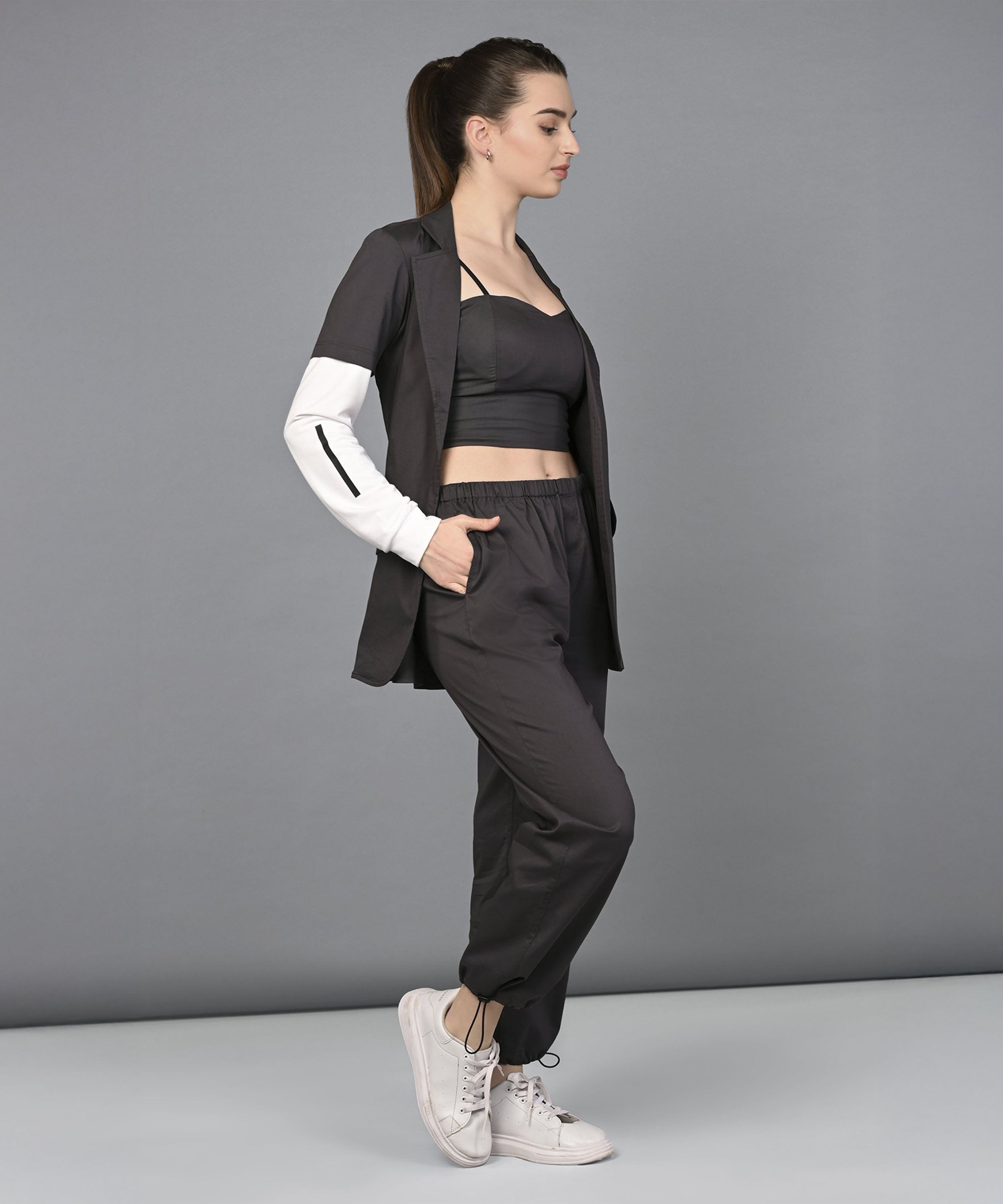 A woman wearing the Athleisure Blazer Tracks Two Piece Co-ord set in black cotton with white sneakers stands sideways against a plain grey background.