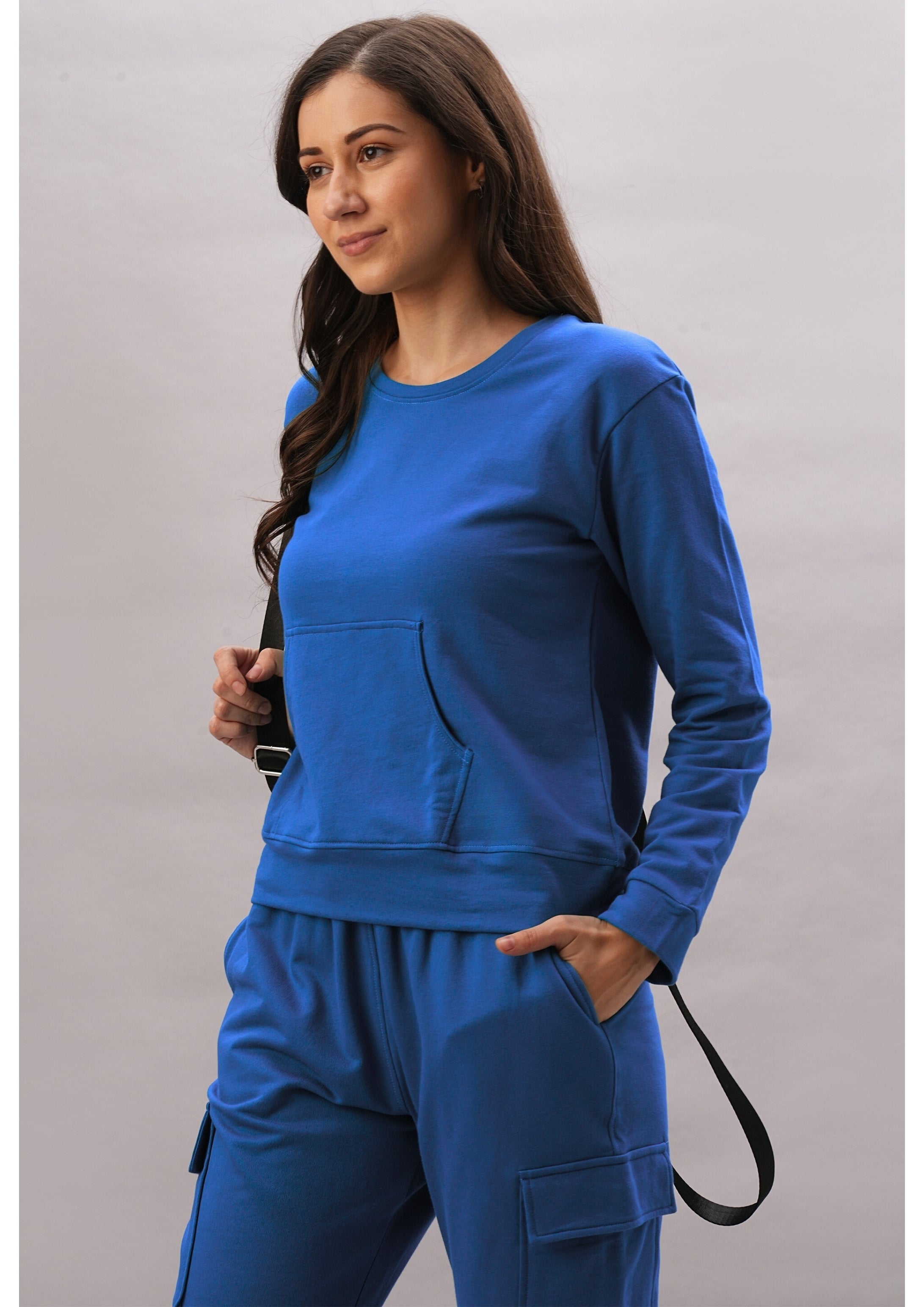 A woman wears the Soft Royal Blue Trackset with cargo-style joggers, her hand in her pocket and holding a black strap, set against a light background.