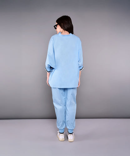 Woman wearing the Denim Accented Set—loose 100% cotton batwing sleeve shirt and joggers—with white sneakers, standing with her back to the camera against a gray backdrop.