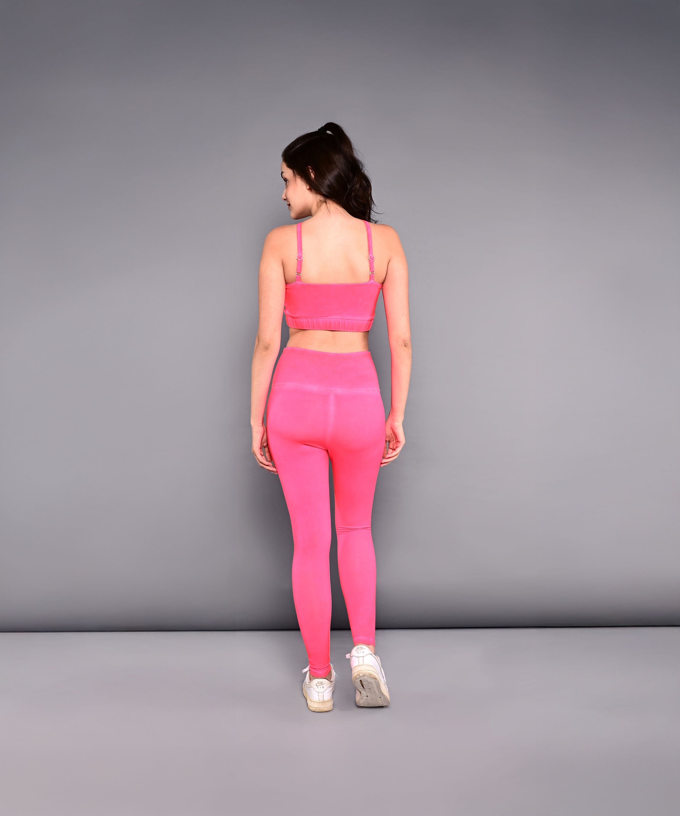 A woman models the Neon Pink Sports Bra, made from skin-friendly cotton lycra, standing with her back to the camera against a plain gray background to highlight its vibrant color and comfortable fit.