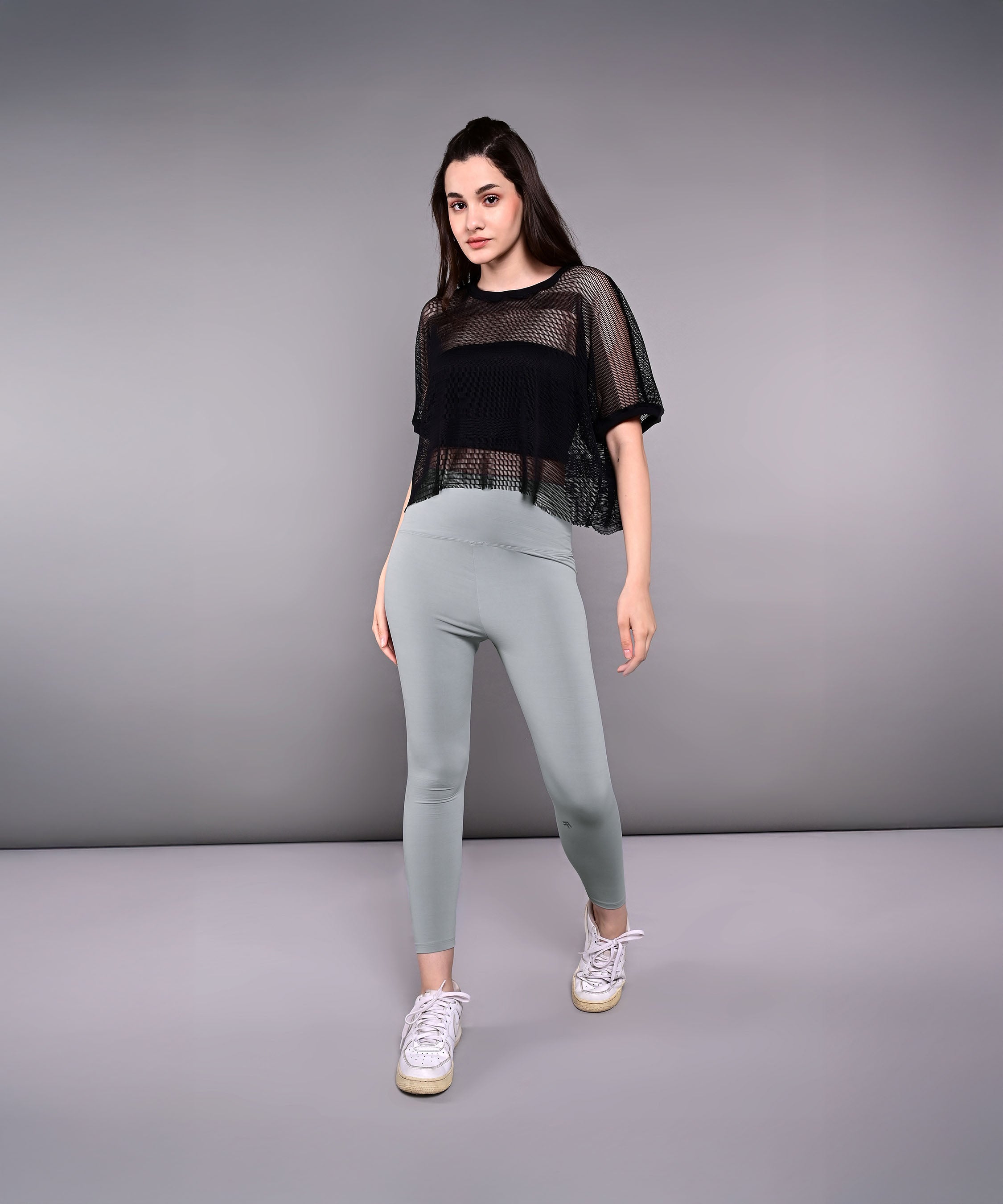 Model wears Tuna Active Black Netted Top, paired with gray leggings and white sneakers, posing against a plain gray background.