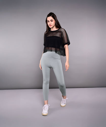 Model wears Tuna Active Black Netted Top, paired with gray leggings and white sneakers, posing against a plain gray background.