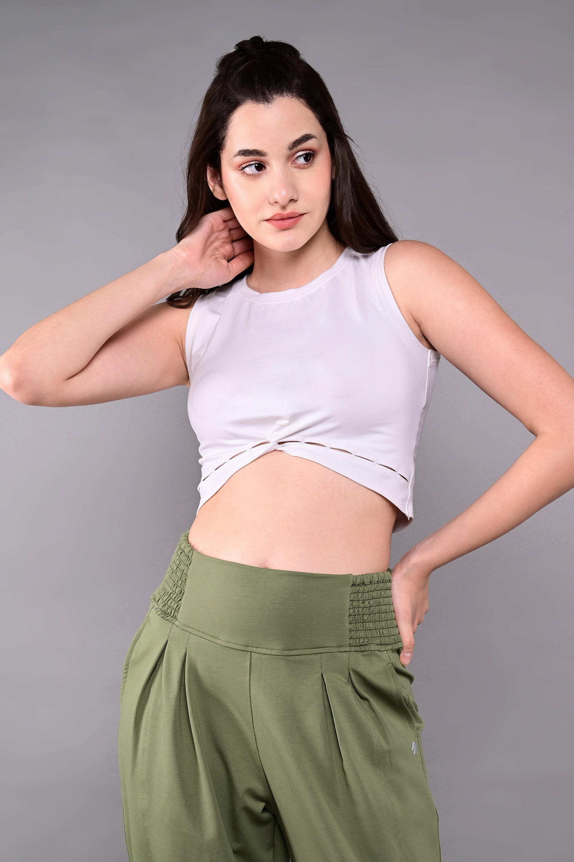 A woman models the White Twisted Crop Top, pairing it with green pants and posing with one hand on her neck against a gray background.