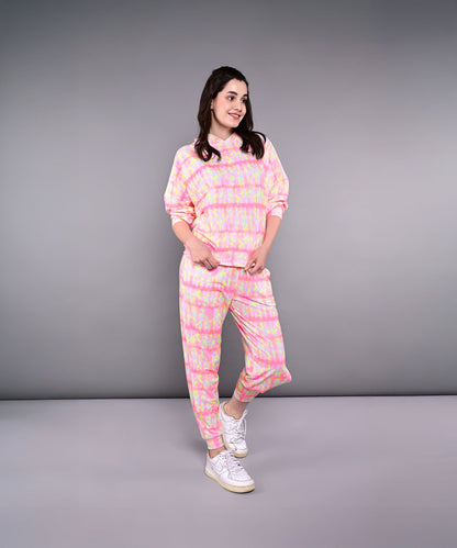 Woman in the Comfort Fit Tie-Dye Sweatshirt With Joggers Set, featuring a pink and yellow tie-dye hooded sweatshirt and joggers in breathable poly-blend fabric, paired with white sneakers, smiling against a gray background.