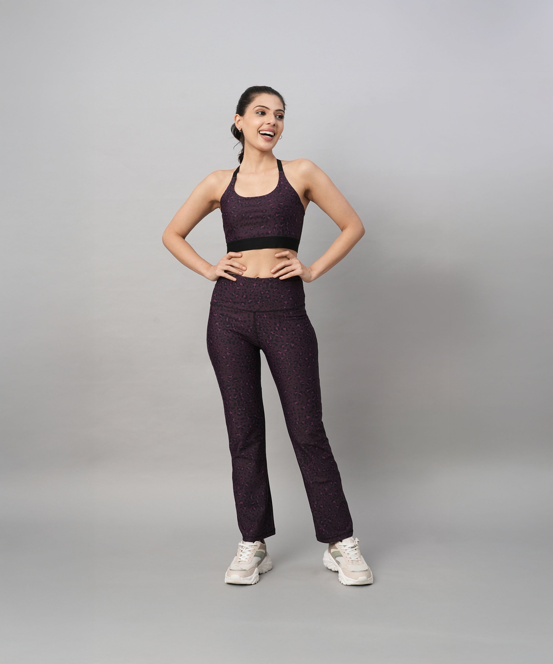 A woman stands smiling against a gray background, wearing stylish activewear: Pink Animal Print Wide Legged Leggings paired with a purple top and white sneakers.