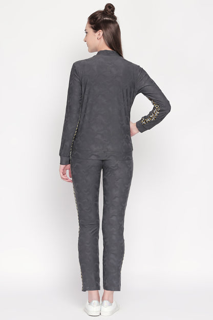A woman stands with her back to the camera, hand on hip, wearing a grey Active Wear Printed Jacket from Tuna London made of comfortable polyester blend, paired with matching bottoms and white shoes.
