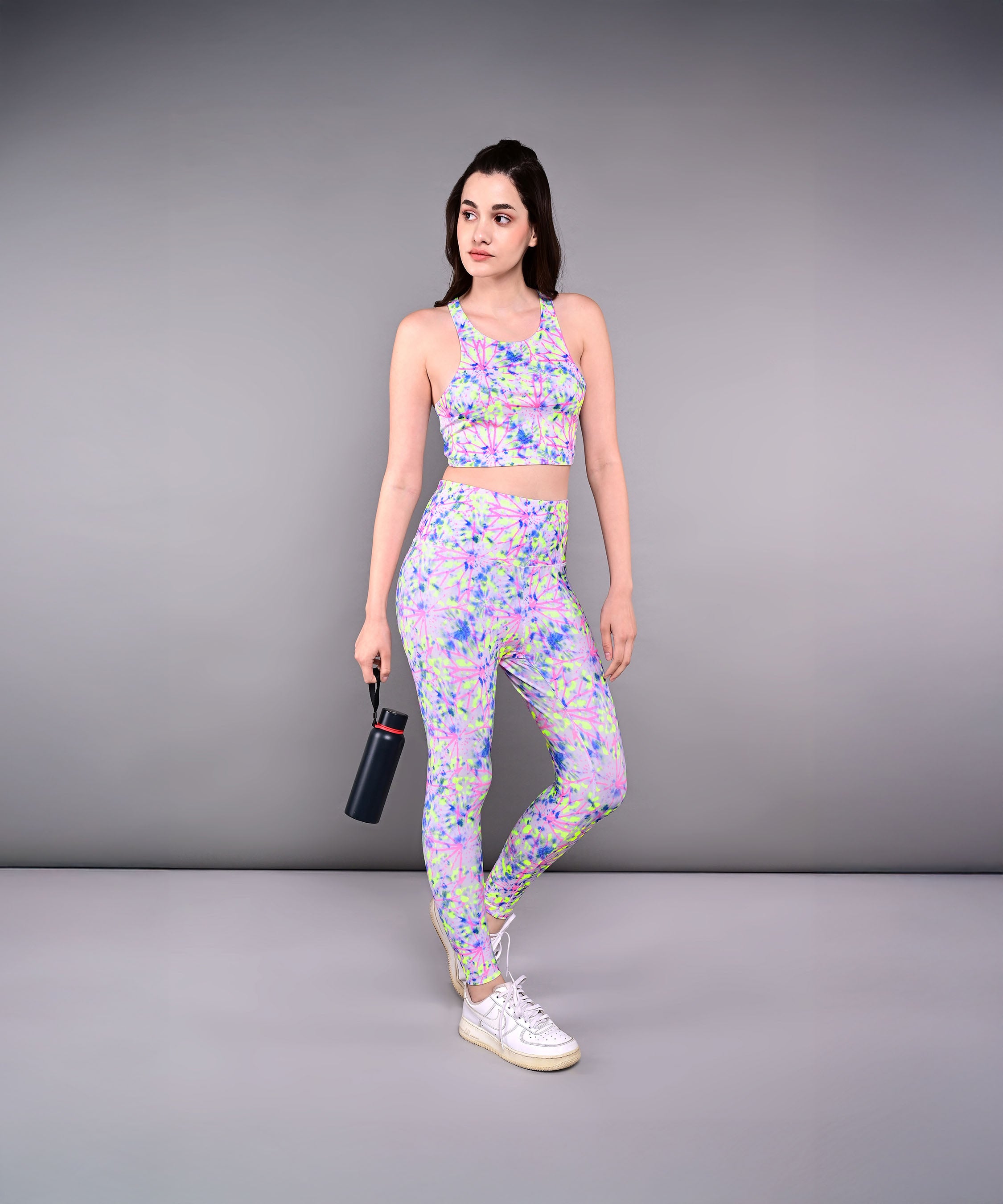 A woman in a colorful workout outfit, featuring Multicolour Neon Leggings and holding a water bottle, stands against a plain gray background.