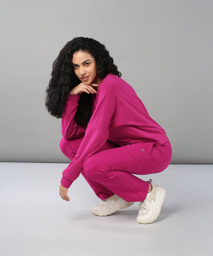 A woman with curly hair squats in the Deep Pink Comfort Duo, pairing a cotton lycra top and matching joggers with white sneakers, set against a gray background.